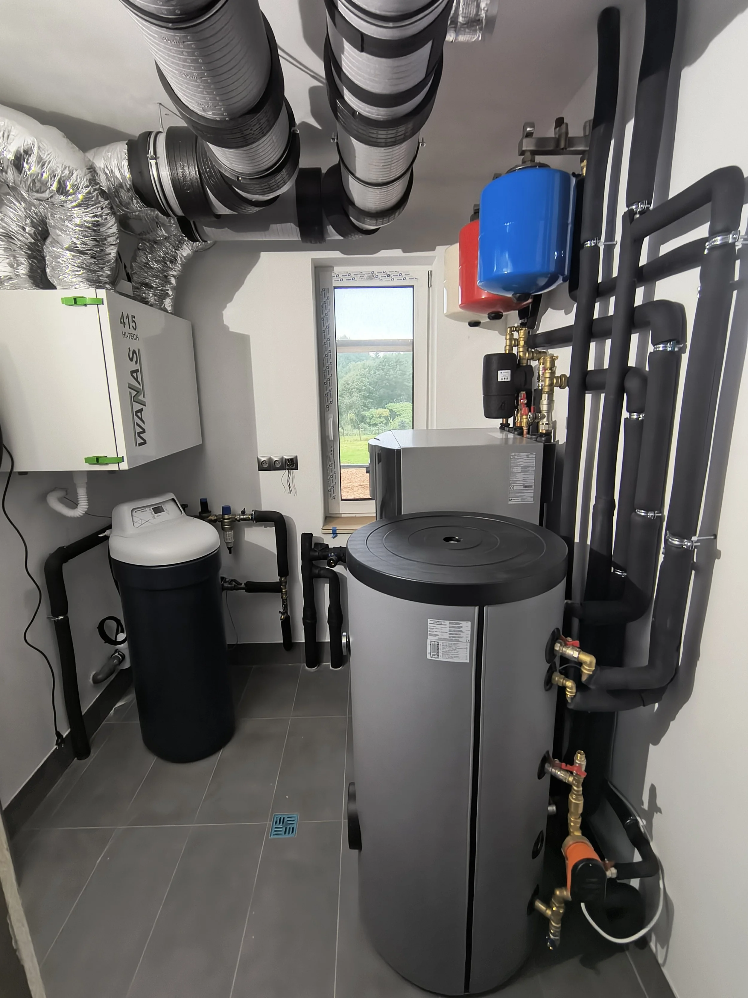A utility room with a water heater, piping, and storage tanks, including a furnace, and a window showing greenery outside.