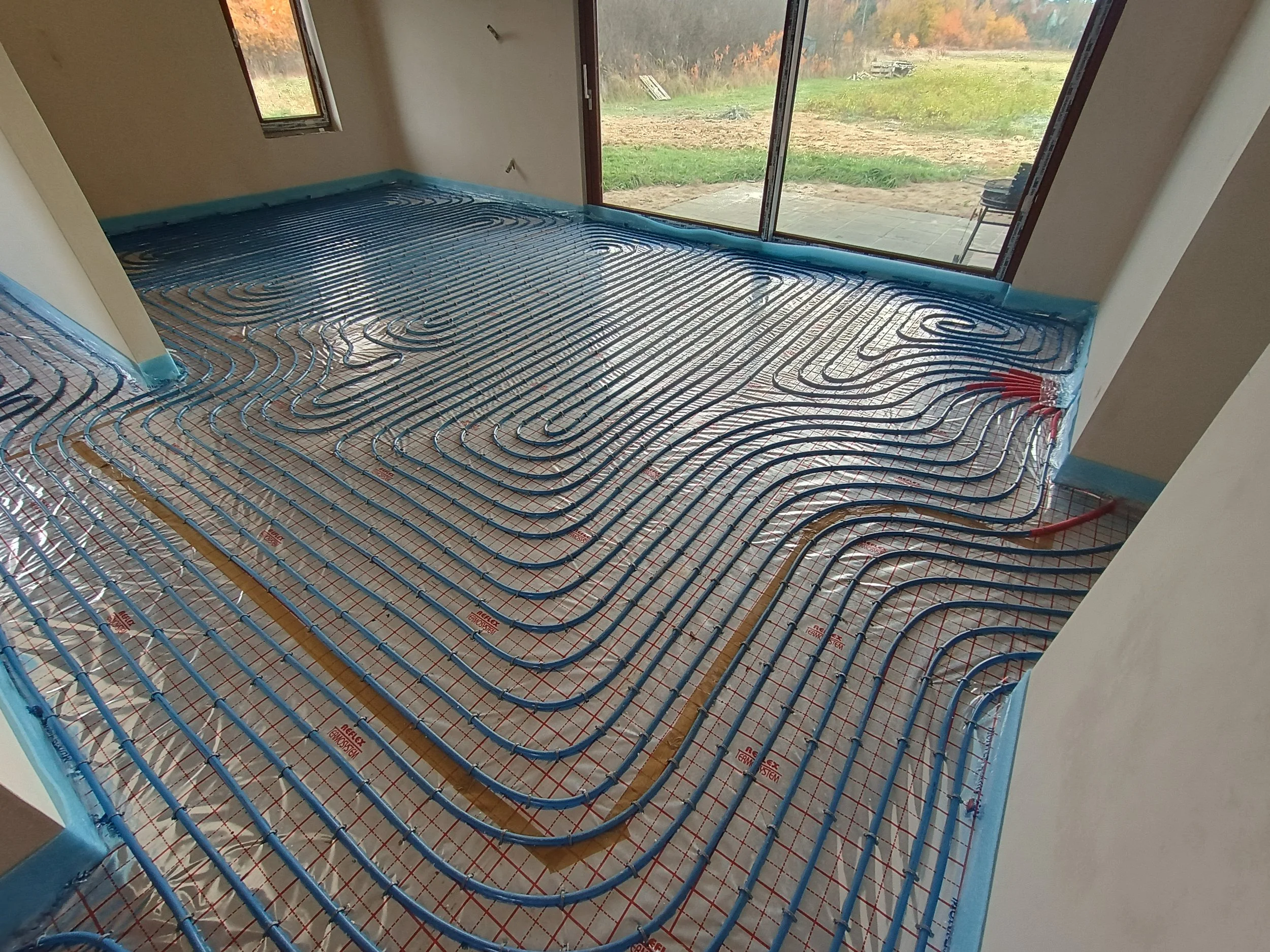 Interior view of a room under construction with radiant floor heating pipes installed on the floor, large windows overlooking a grassy outdoor area, and an open door leading outside.