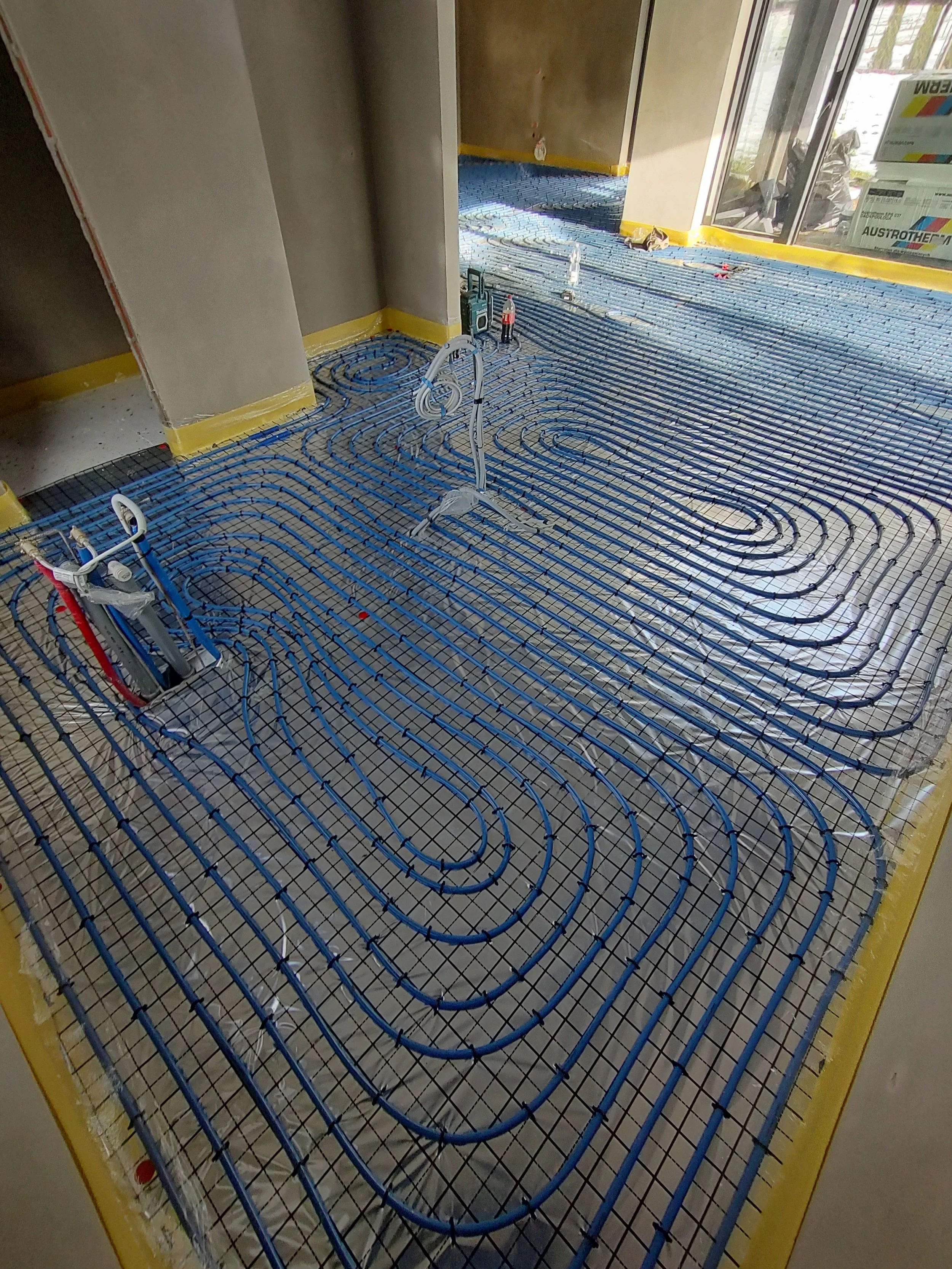 Construction site with radiant floor heating tubes being installed, inside a building with large windows.