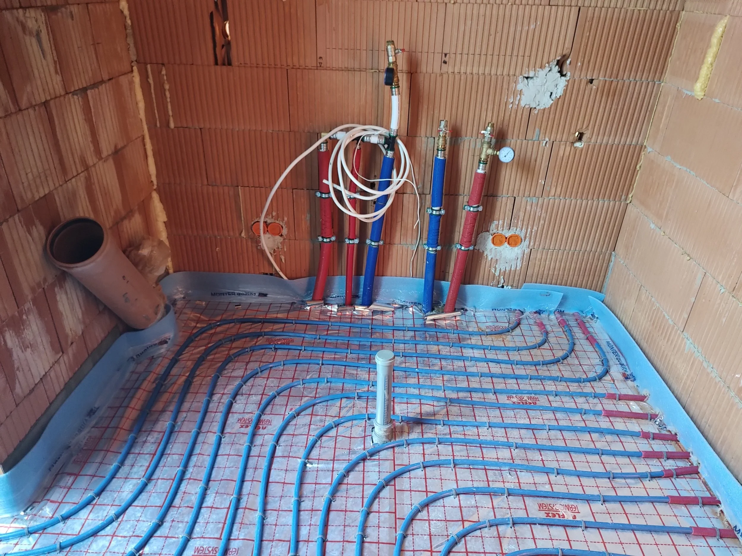 Bathroom under construction with plumbing pipes installed, insulation, and a drain pipe on the wall with exposed brick walls.