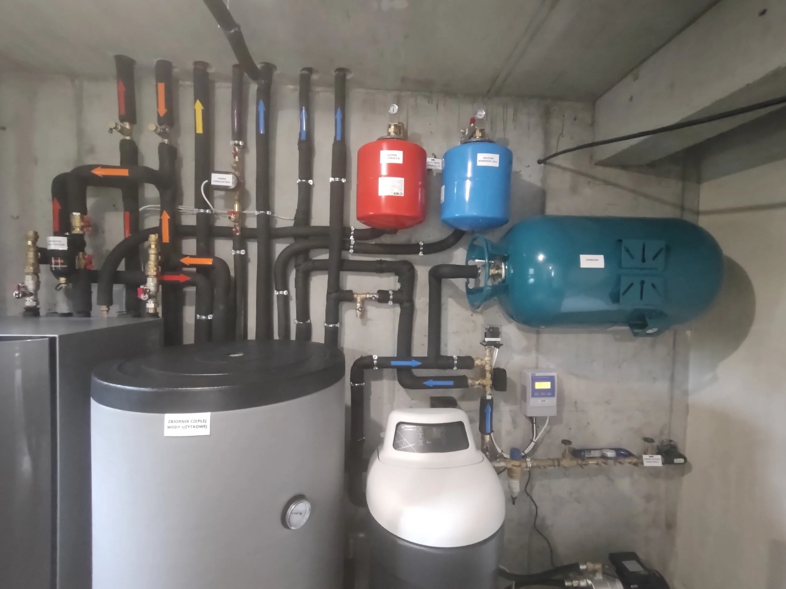 A utility room with black pipes, red and blue expansion tanks, a water softener, and a white water heater. The pipes are labeled with arrows in red, orange, yellow, and blue, indicating water flow directions.