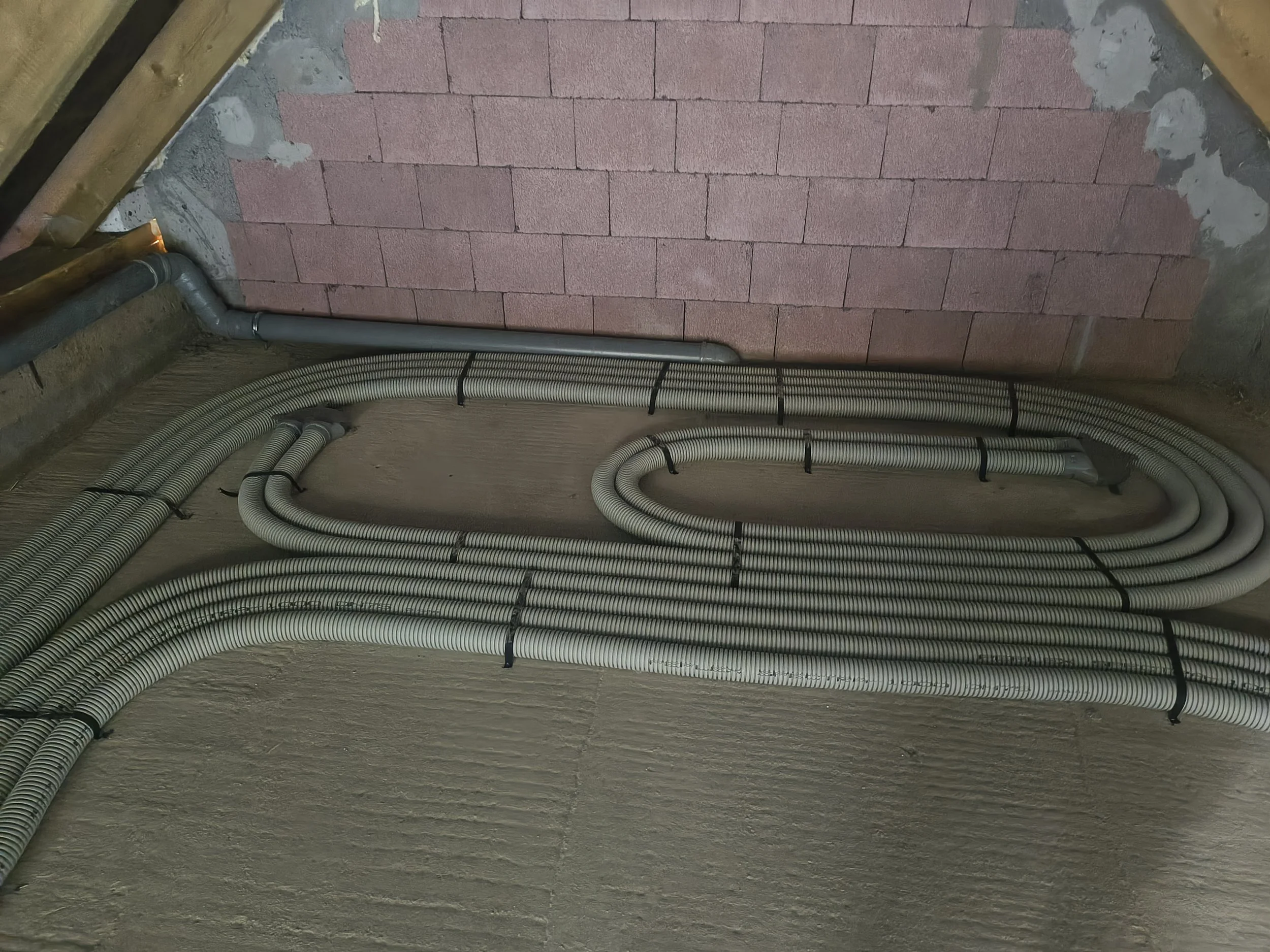 A wall under construction with pink concrete bricks and grey mortar. Several black zip ties secure grey and white flexible conduit pipes to the wall, arranged in concentric curves at the bottom of the image. Parts of wooden framing and a metal pipe a