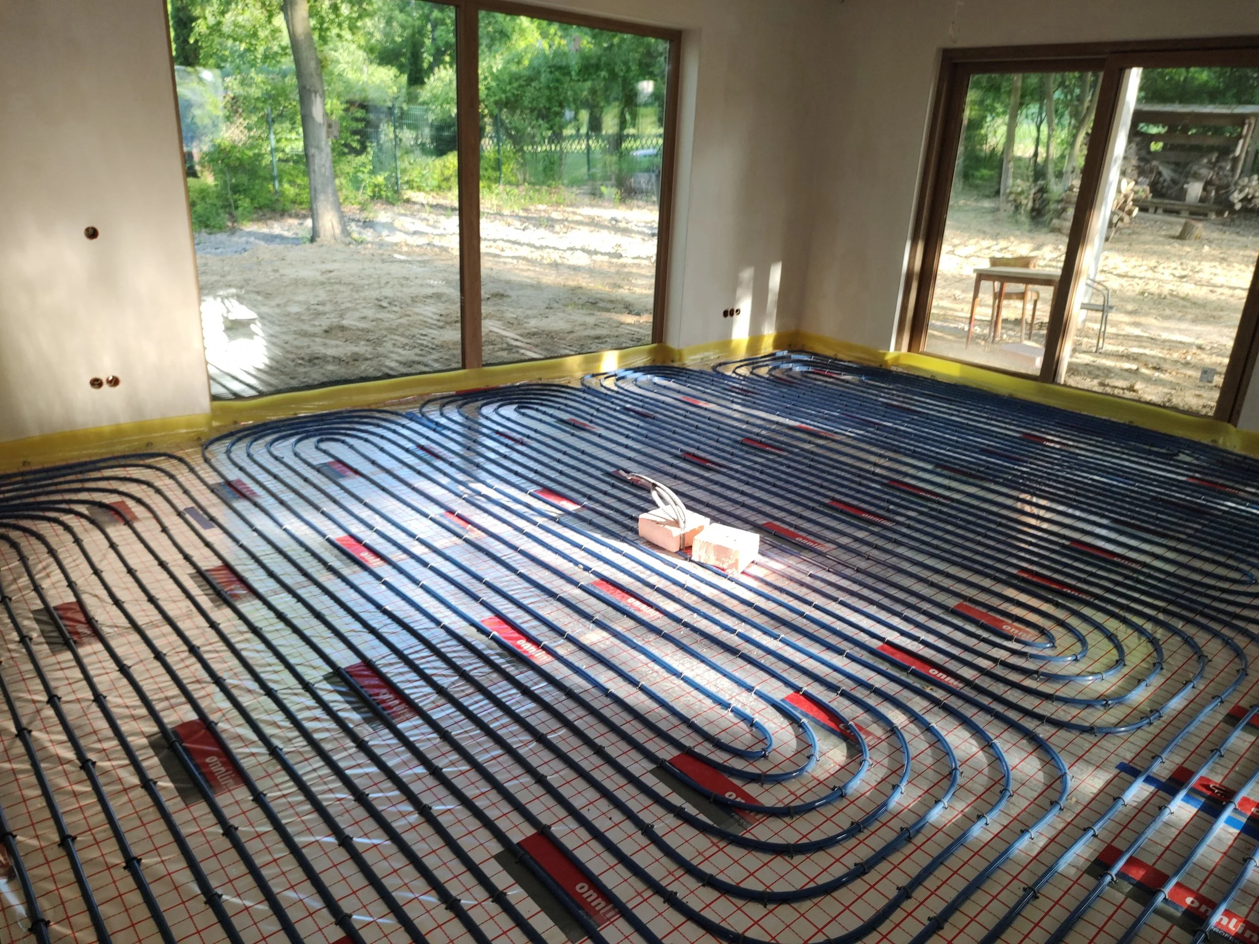 Interior of a house under construction, showing radiant floor heating with black heater tubes installed on a red and white grid, ready for concrete pouring, with large windows overlooking an outdoor yard and trees.