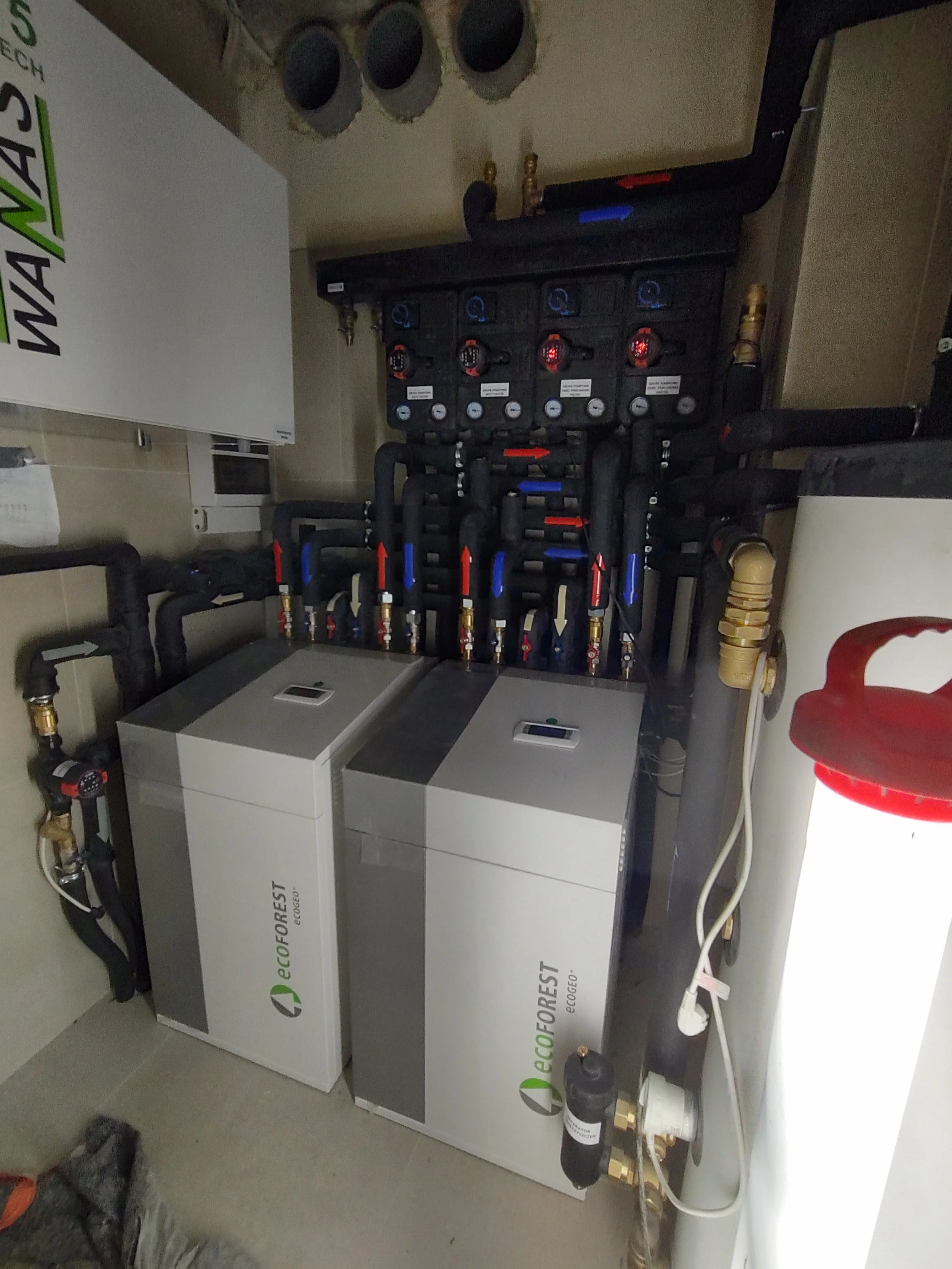 An indoor plumbing system with black piping, gauges, and white units labeled 'ecoFOREST'. There are also brass fittings, a red chair, and a fire extinguisher nearby.