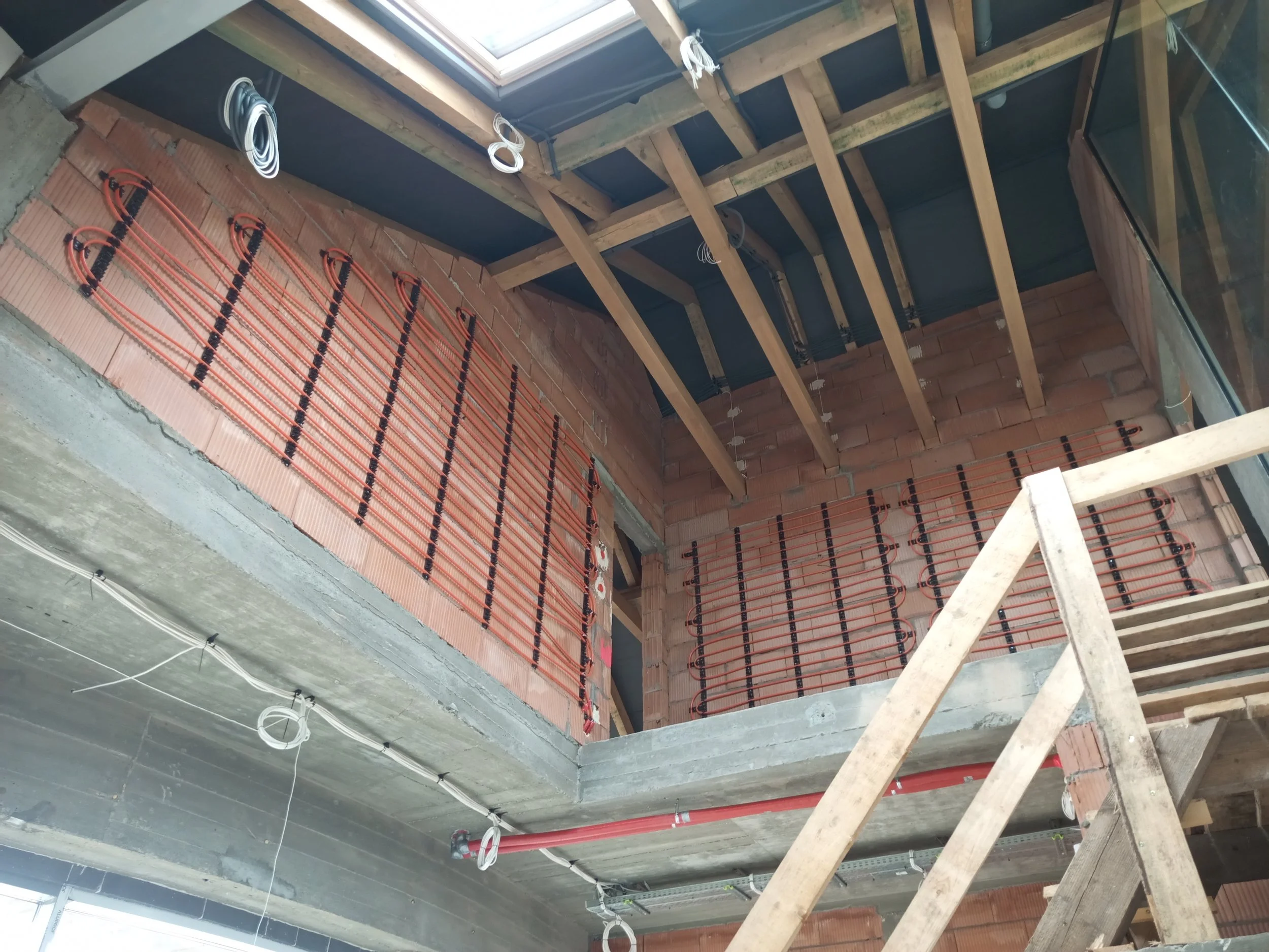 Interior view of a building under construction, showing exposed brick walls, ceiling framework with wooden beams, electrical wiring, black and orange tubing for radiant heating, and a skylight window.
