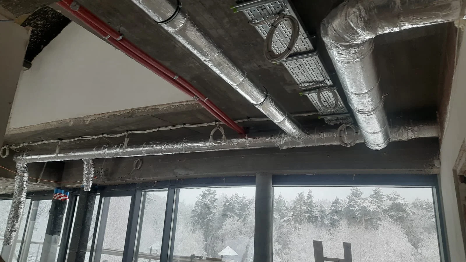 Construction site ceiling with exposed ductwork, electrical wiring, and window showing snowy trees outside.
