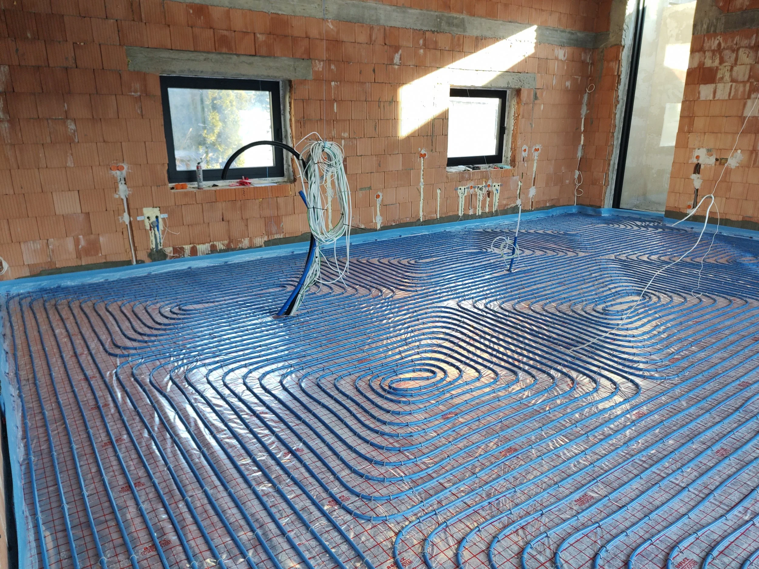 Interior of a building under construction with exposed brick walls, three windows, and a floor heating system being installed with blue tubing laid out in a spiral pattern.