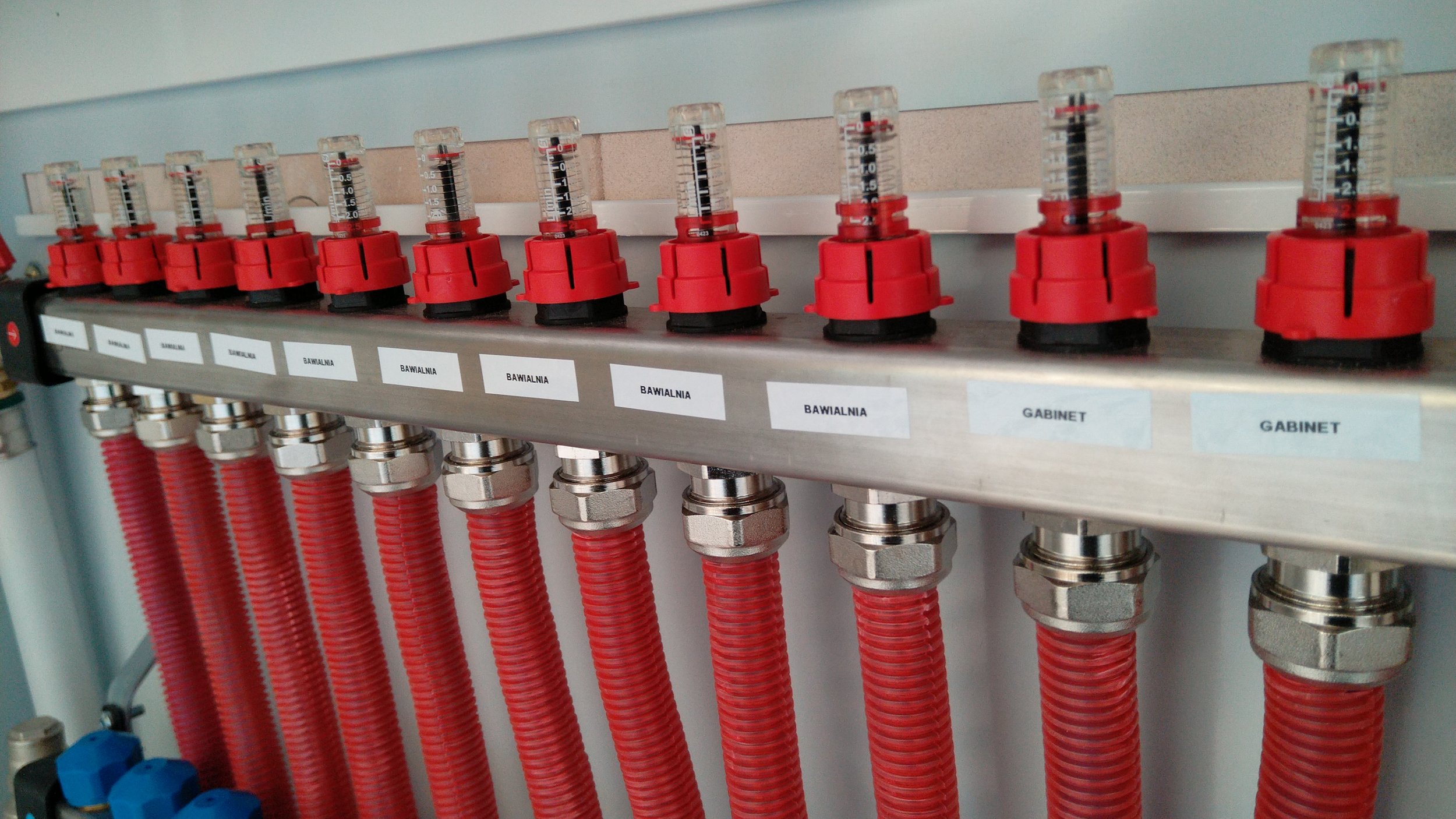 An equipment panel with several red valve handles with attached clear flow meters labeled in Polish, such as 'Bawiarnia' and 'Gabinet', connected to red hoses and metallic fittings.