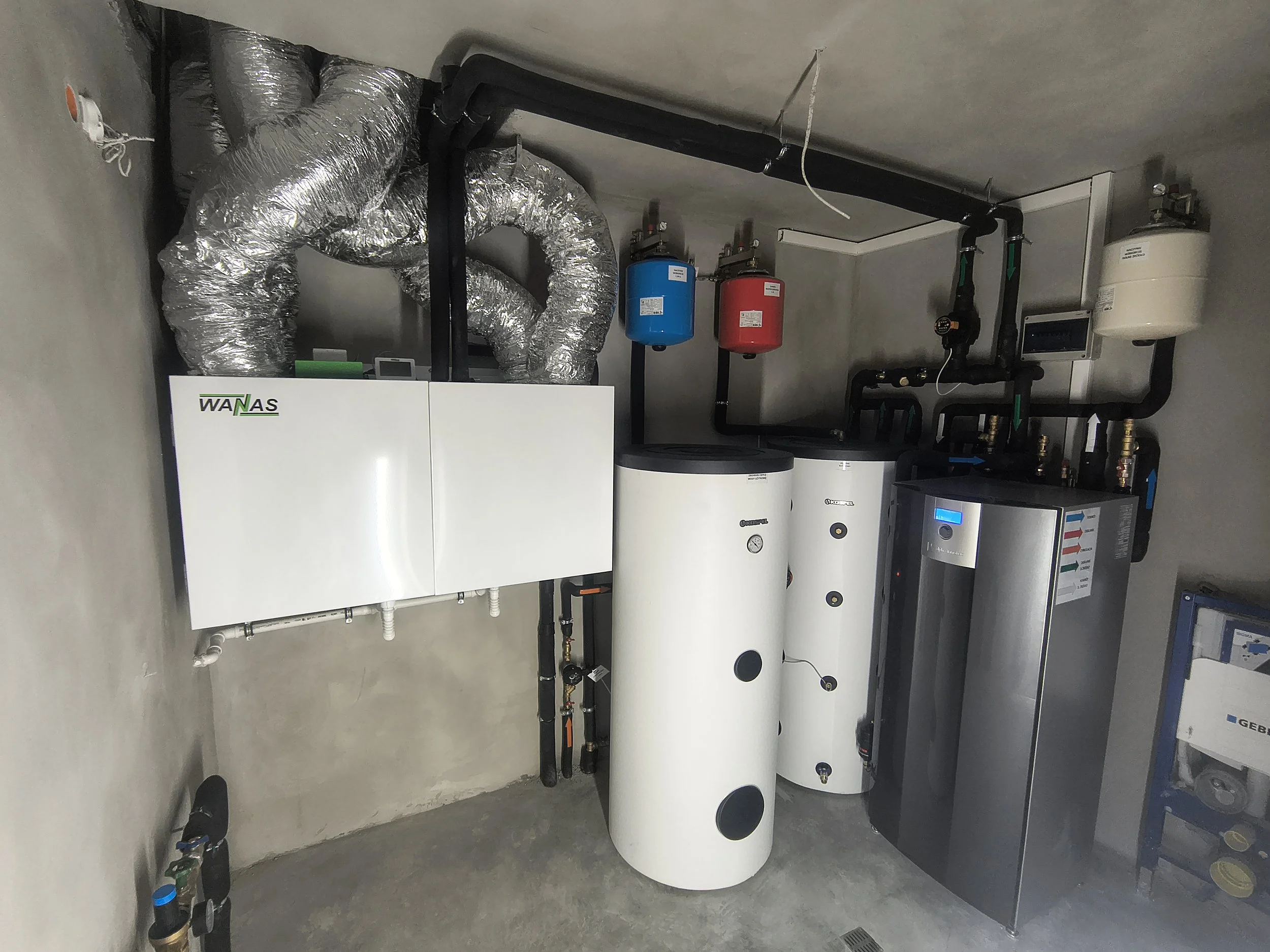A mechanical room with insulated pipes, two water heaters, and control units. The pipes run along the ceiling and walls, with some connected to blue and red expansion tanks.