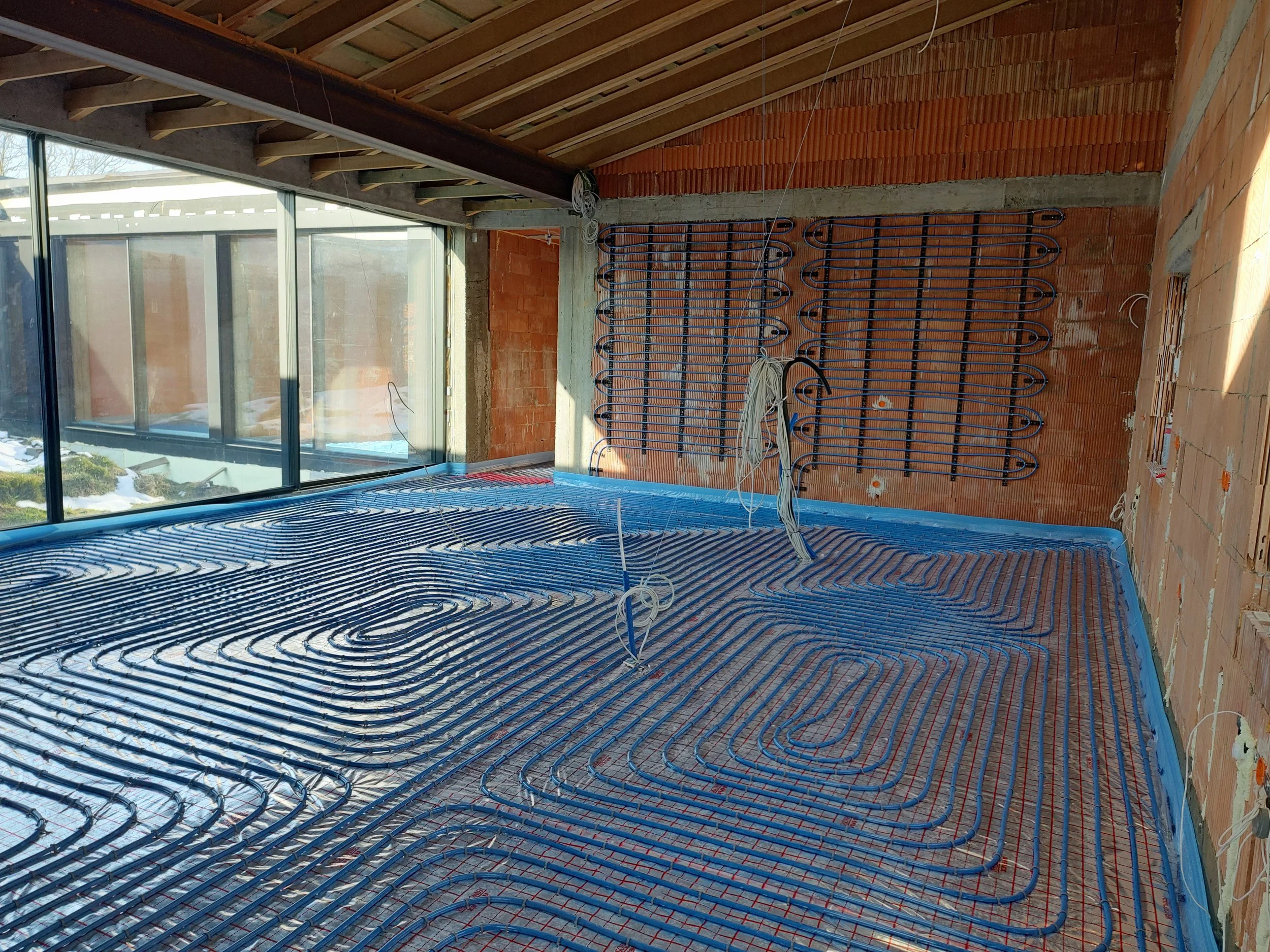Interior of a building under construction with radiant floor heating system installed, showing blue tubing on the floor, red wiring, and unfinished brick and wood structure.