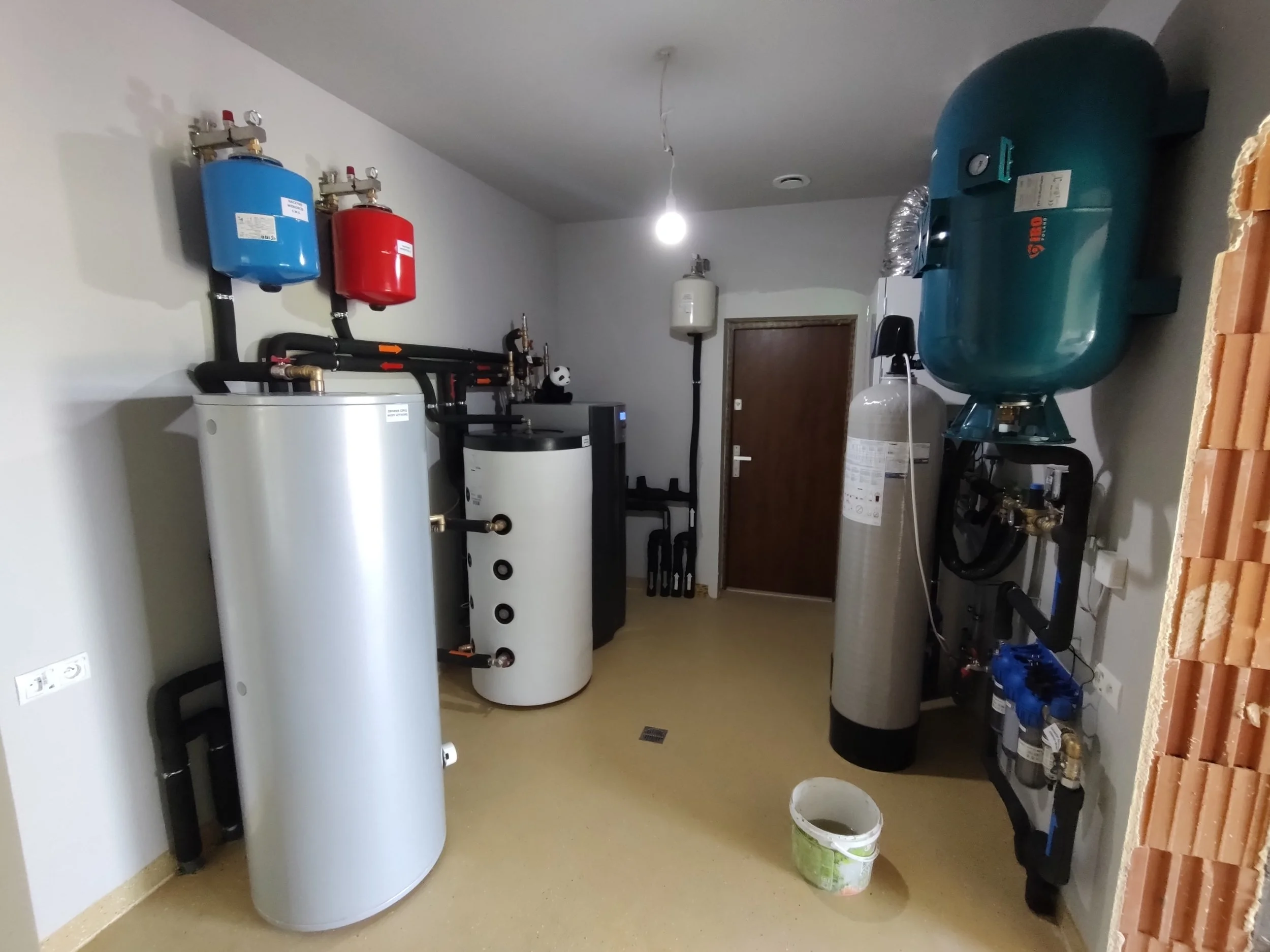 A utility room with a water heater, a large air compressor, and various plumbing and heating components. The room has beige flooring, white walls, and a partially exposed brick wall on the right.