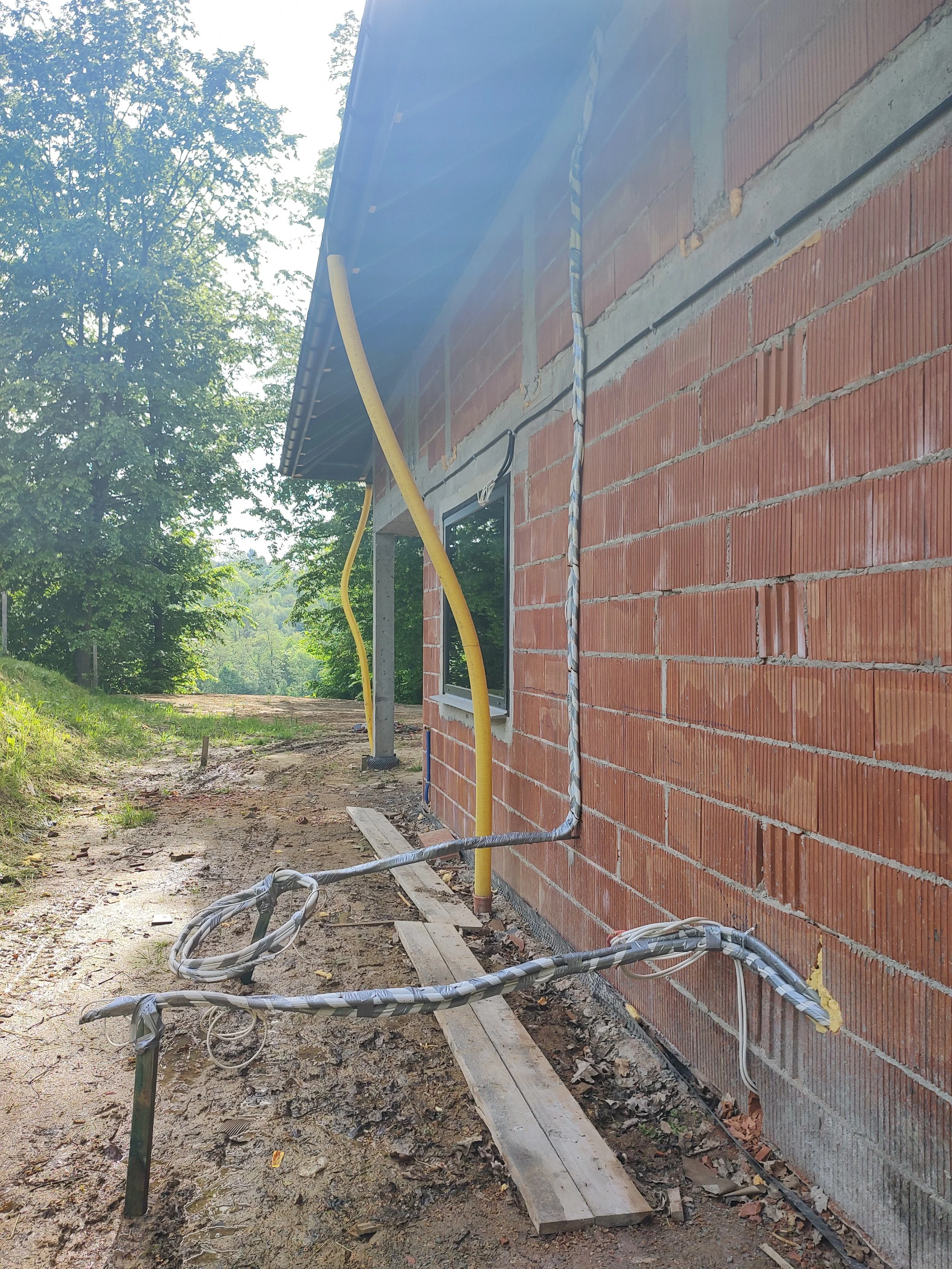 Construction site showing the exterior of a brick building with electrical conduit and cable work in progress, surrounded by dirt and construction materials.