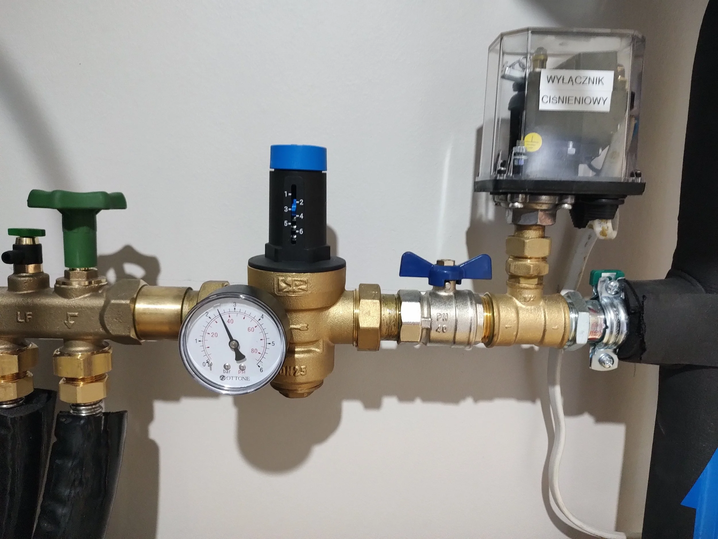 A plumbing setup with brass valves, a pressure gauge, and a pressure switch, with stops labeled 'LF' and 'F' and a switch labeled 'WYŁĄCZNIK CIŚNIENIOWY' mounted on a white wall.