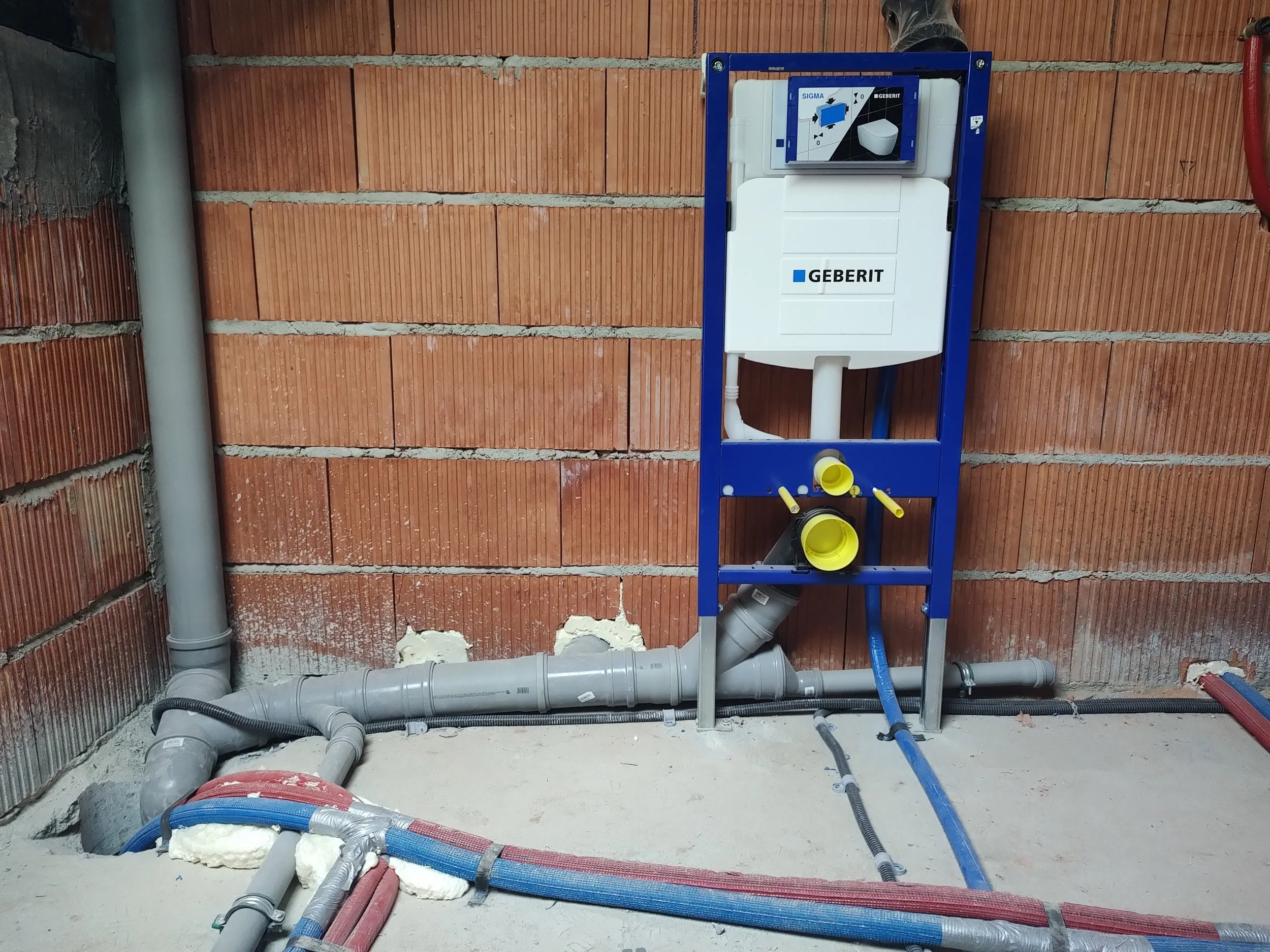 Installation of a toilet flush tank with plumbing pipes, a brick wall background, and construction materials on the floor.