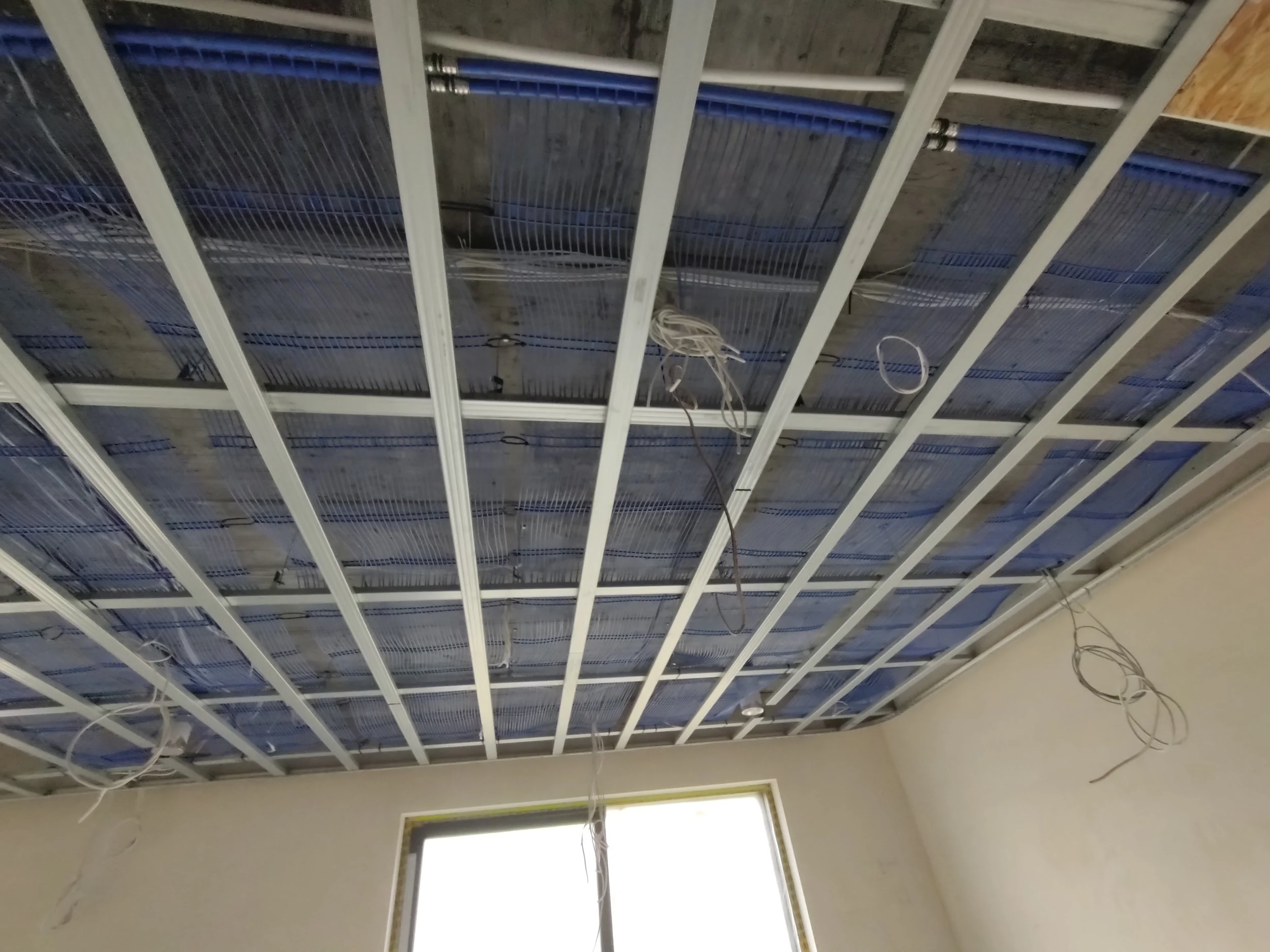 Ceiling under construction with electrical wiring and metal framing for ceiling panels, with a window below letting in natural light.