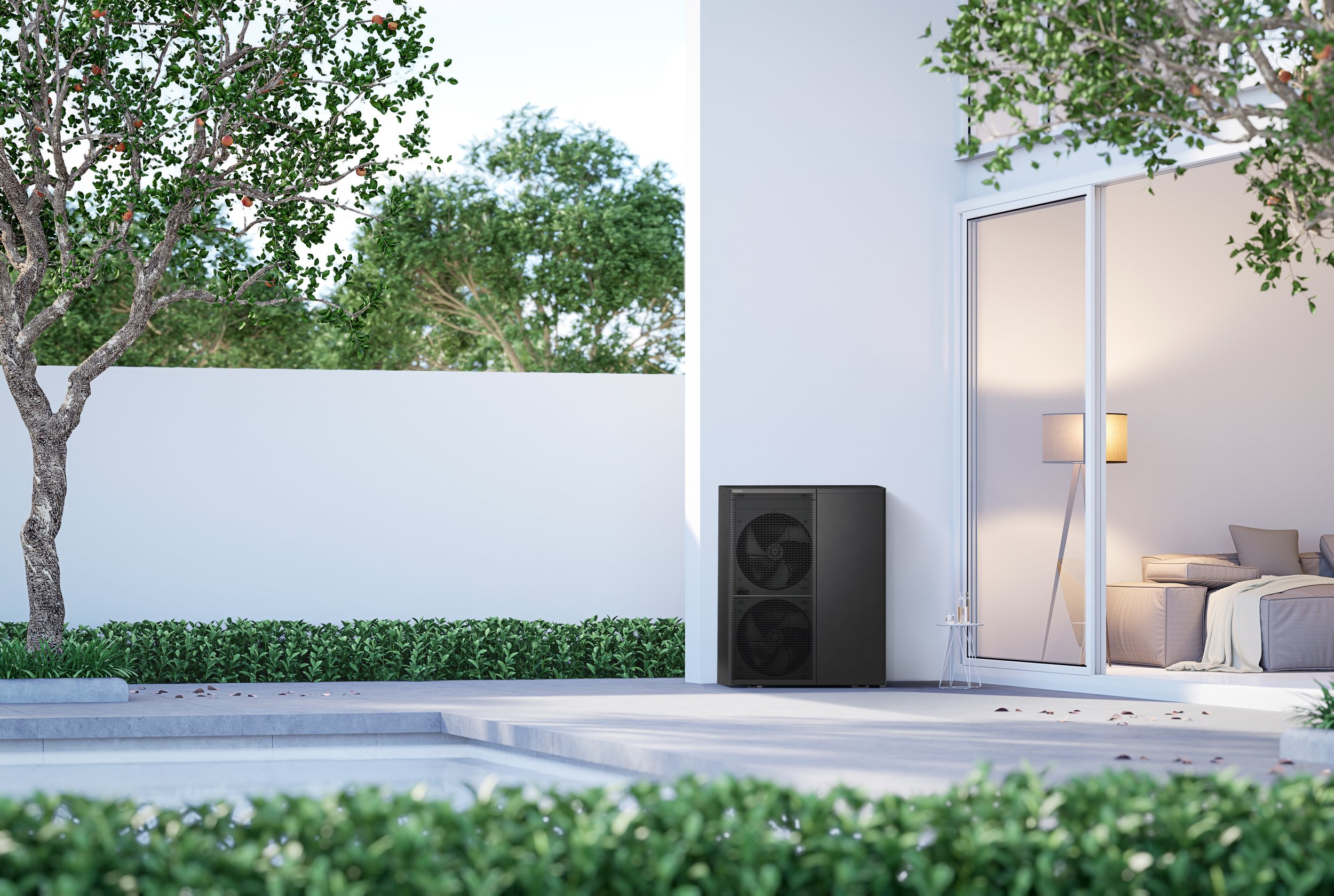 Modern house patio with a white exterior wall, a large glass sliding door showing a cozy interior with a sofa and lamp. There is a tall black speaker or planter near the door. The patio has a layered design with steps, surrounded by green shrubs and trees, including a pomegranate tree with red fruit.