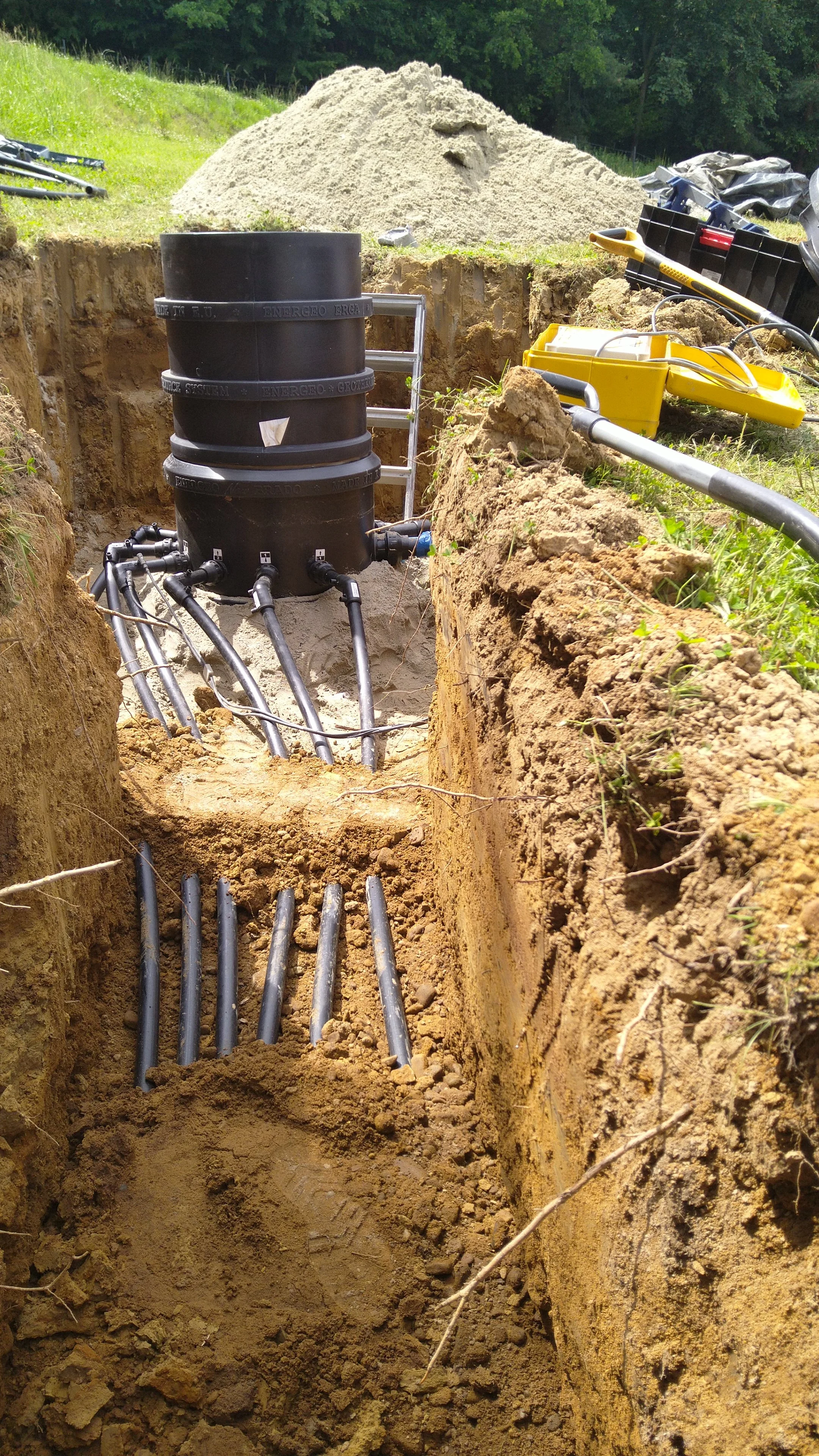 An underground utility installation with black pipes connected to a black cylindrical structure in a trench, surrounded by dirt, with construction tools nearby and a mound of dirt in the background.