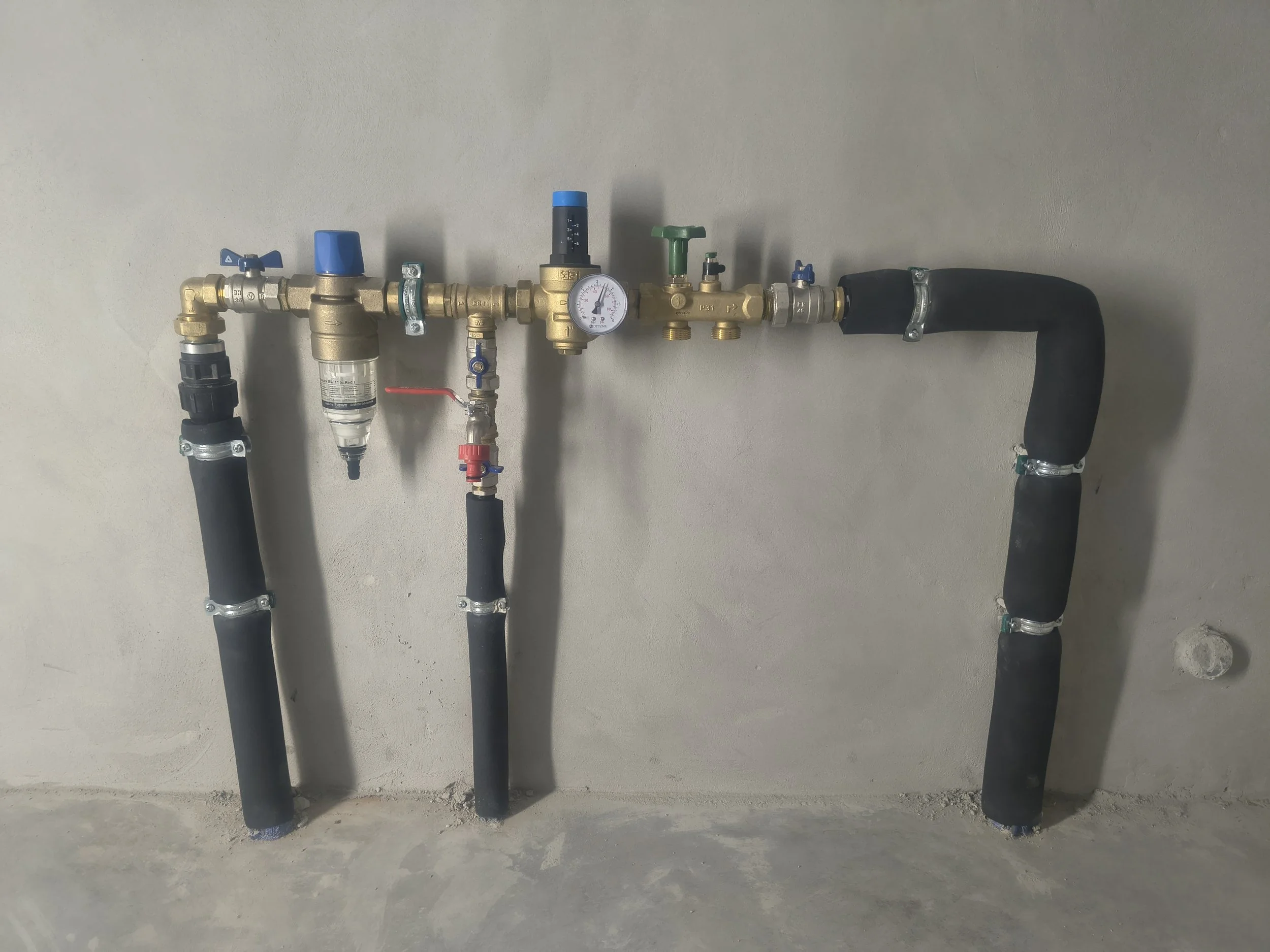 A wall-mounted plumbing system with multiple brass pipes, valves, gauges, and black insulated pipes against a plain wall.