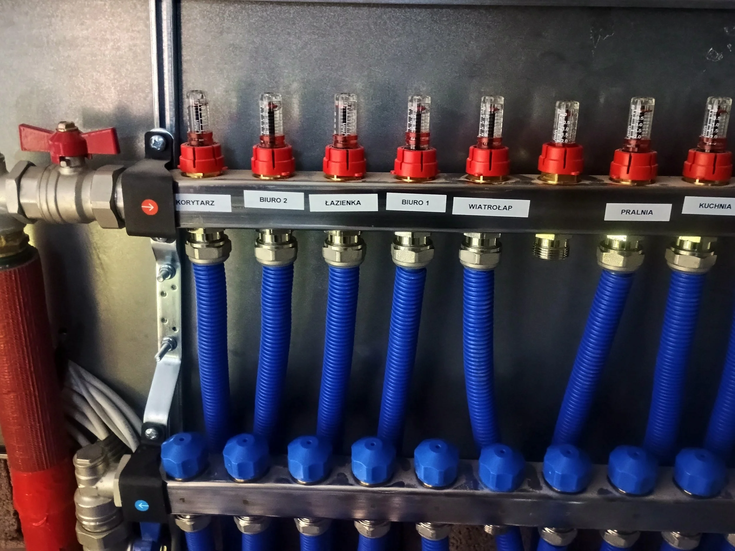 A plumbing manifold with multiple blue hoses connected, labeled with room names in Polish, including corridor, office 2, bathroom, office 1, windbreak, laundry, and kitchen.