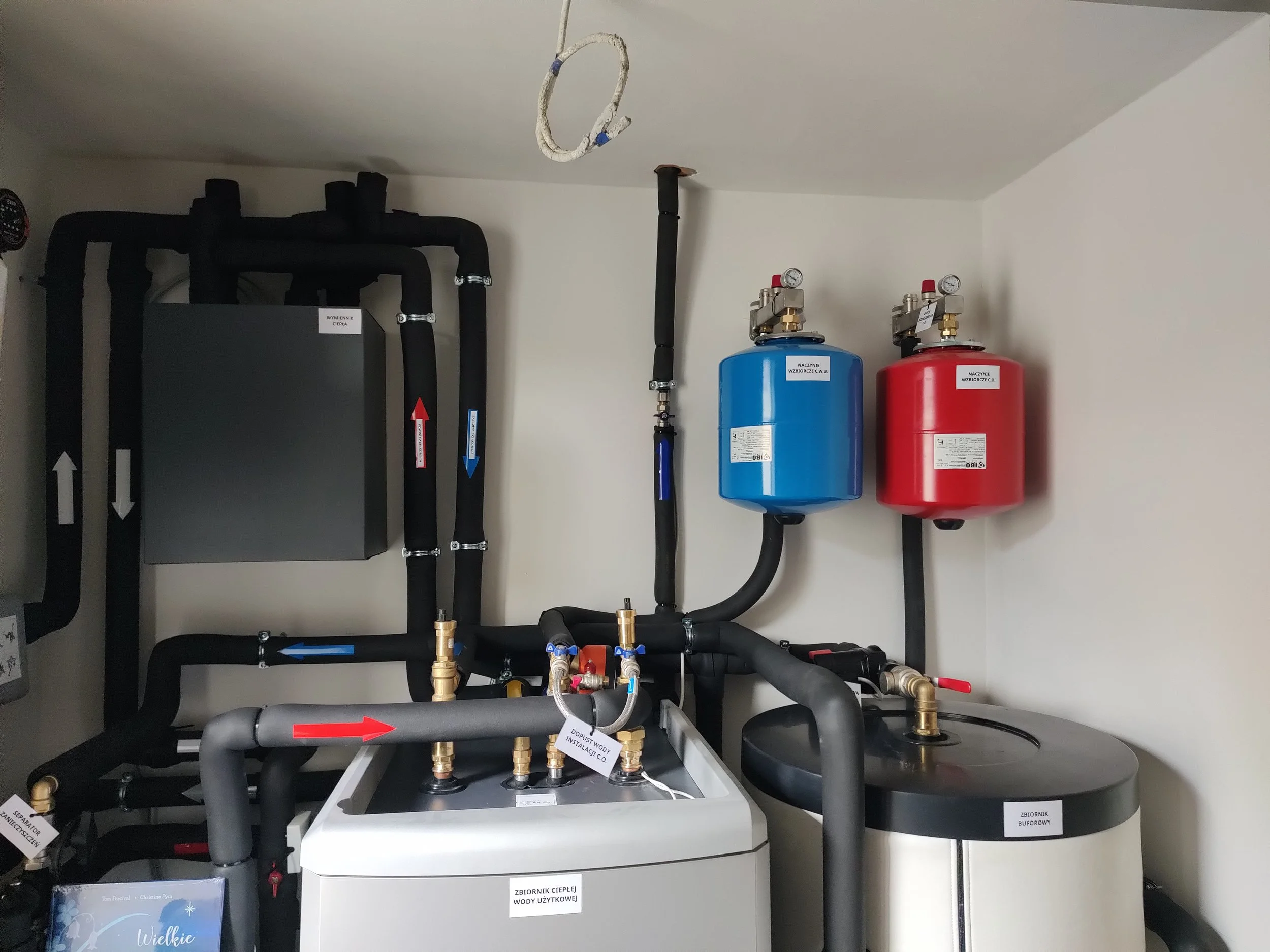 A utility room with black pipes, a gray equipment box, a blue and a red expansion tank, and several valves and gauges.