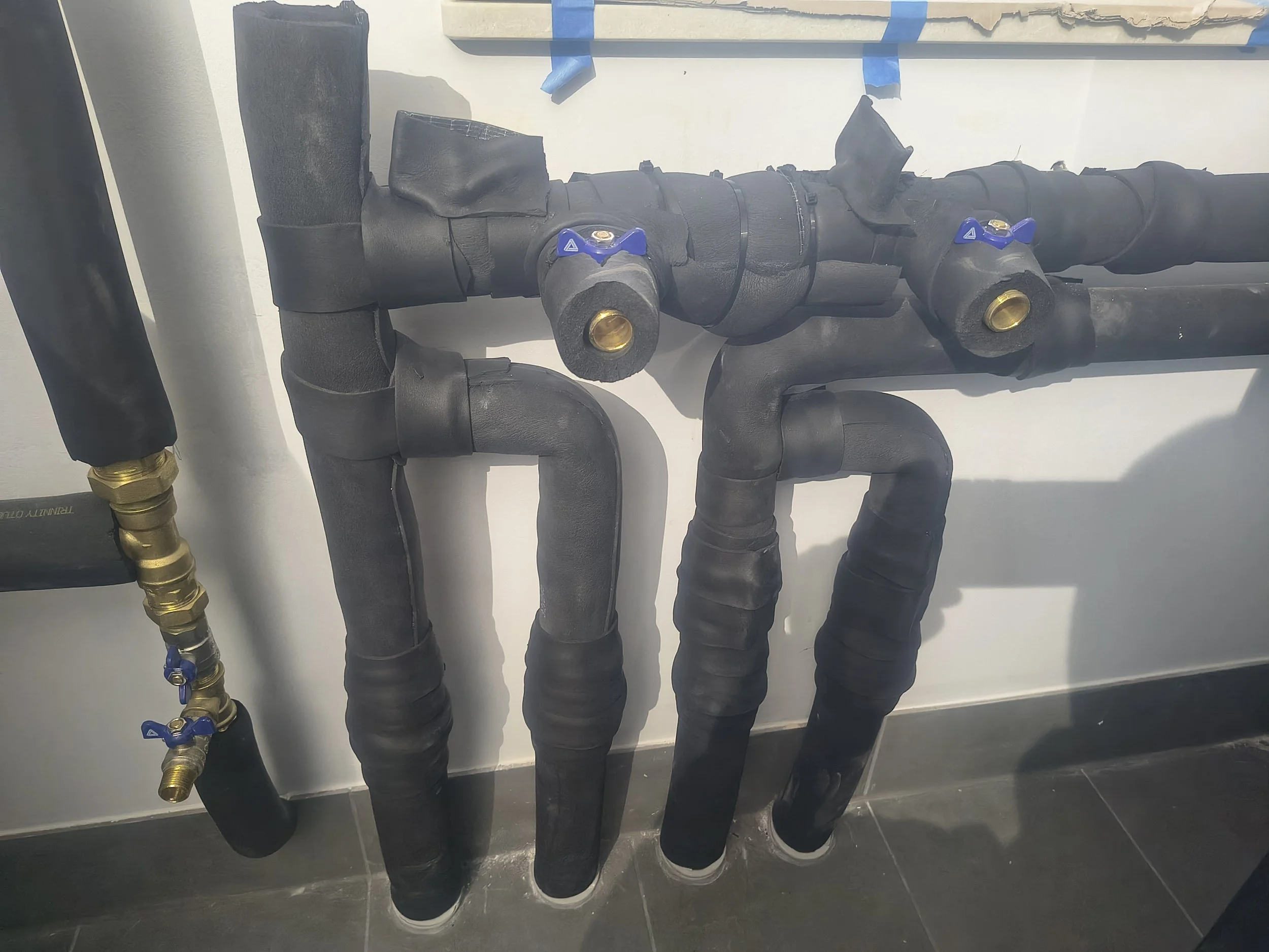 A bathroom wall with black plastic pipes and blue valve handles, connected to brass fittings and a black pipe, mounted on a white wall with some blue tape.