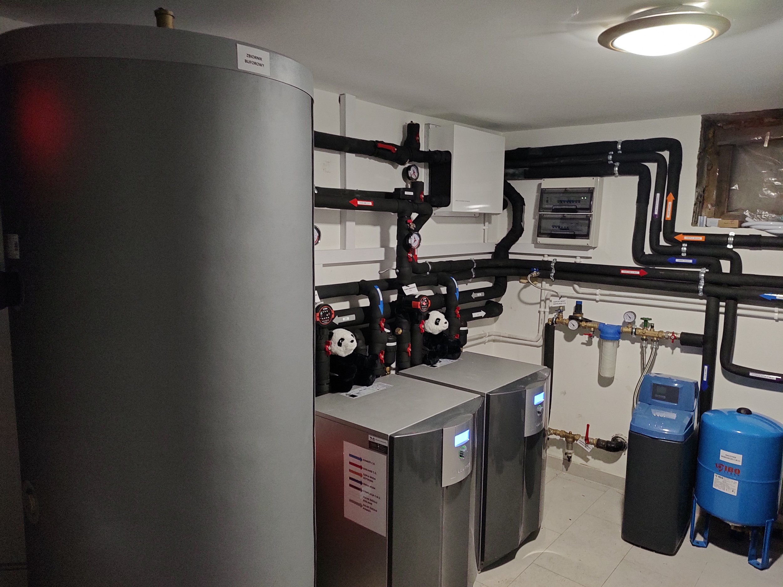 A utility room with black piping, gauges, and various equipment, including a blue water filter and a gray cylindrical appliance.