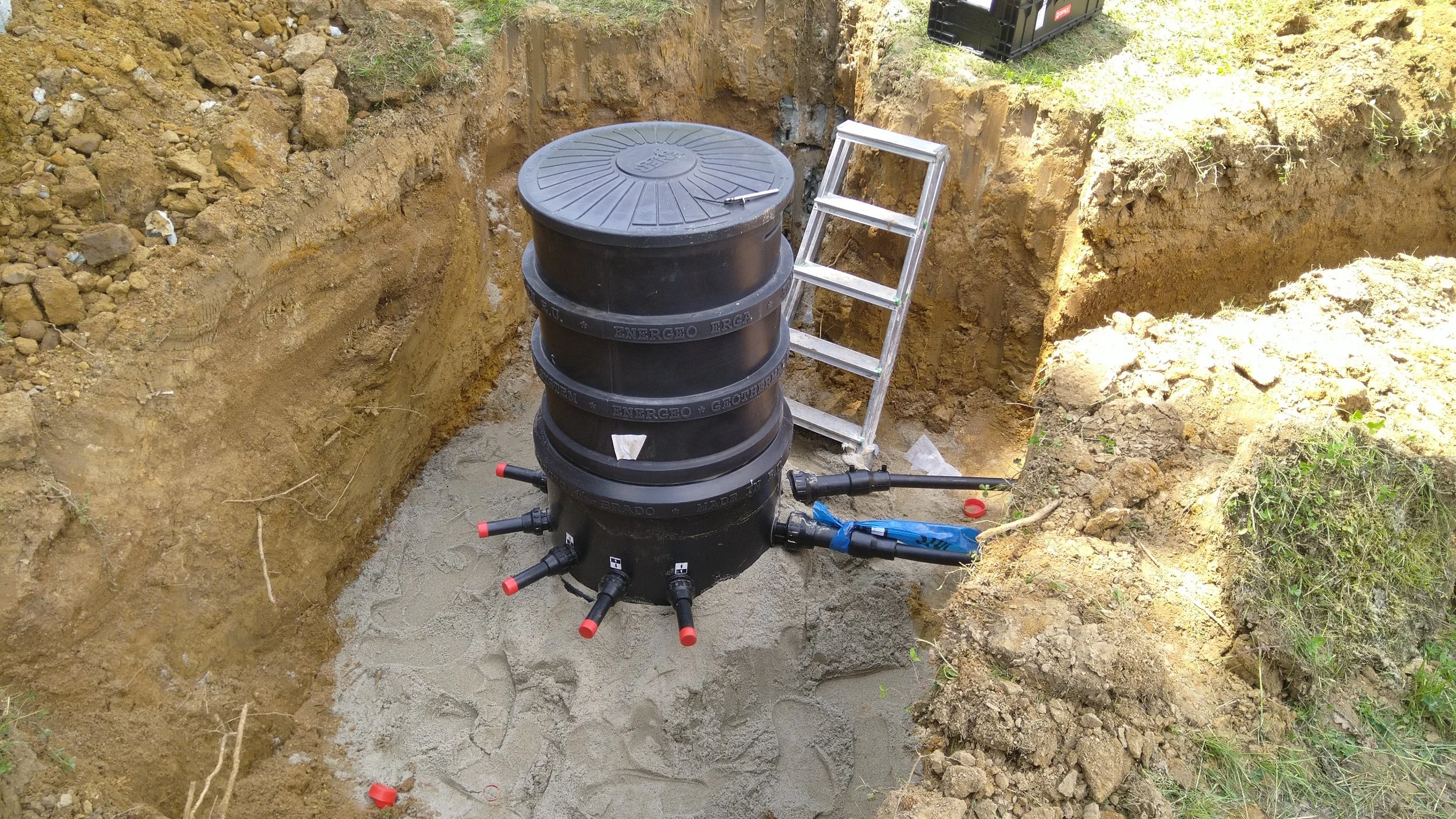 A large underground water storage tank being installed in a trench, with a small ladder leaning against it and pipes connected at the bottom.