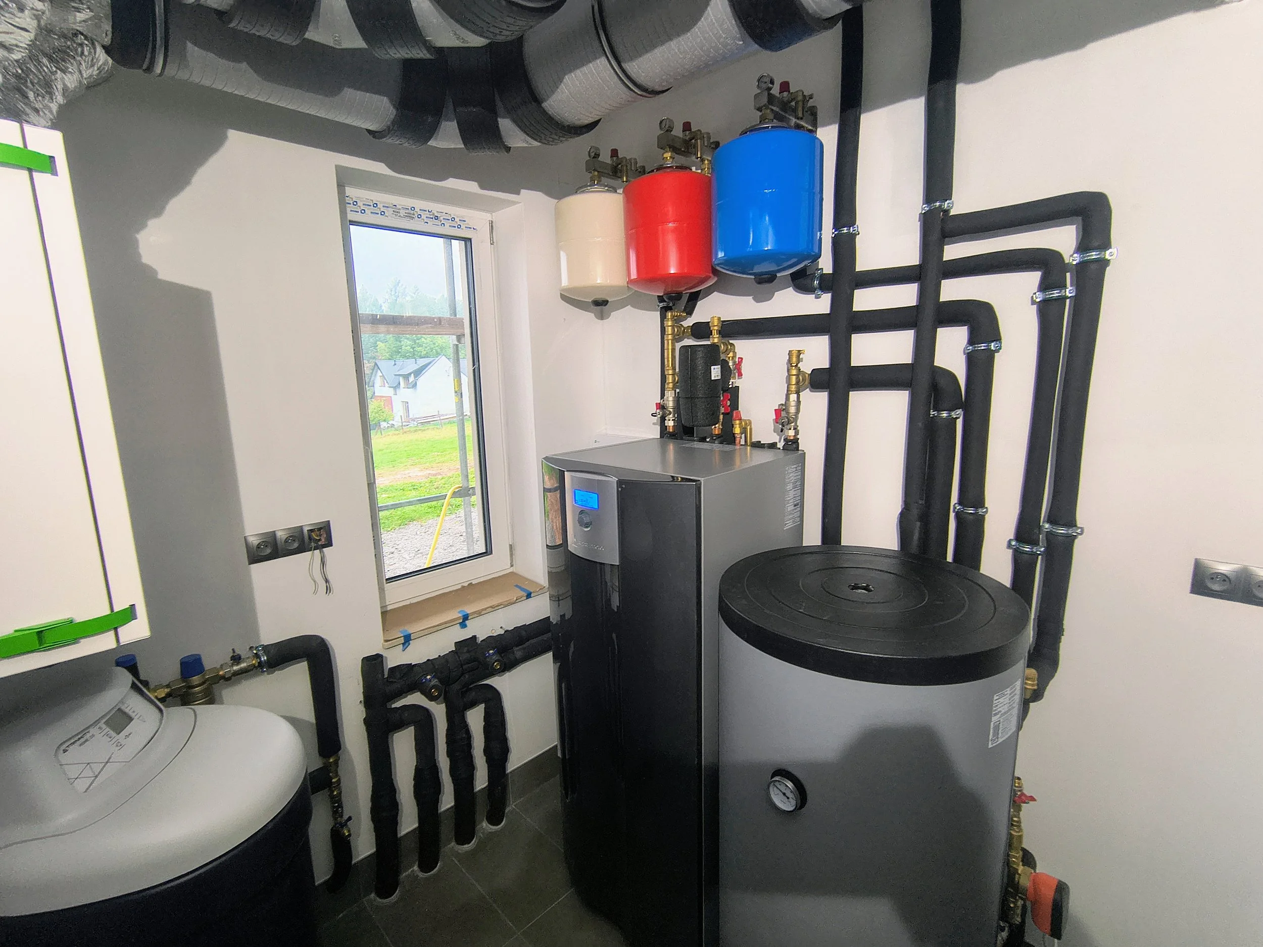 A mechanical room with various pipes, tanks, and a control panel. Outside through the window, there are houses and a green lawn.
