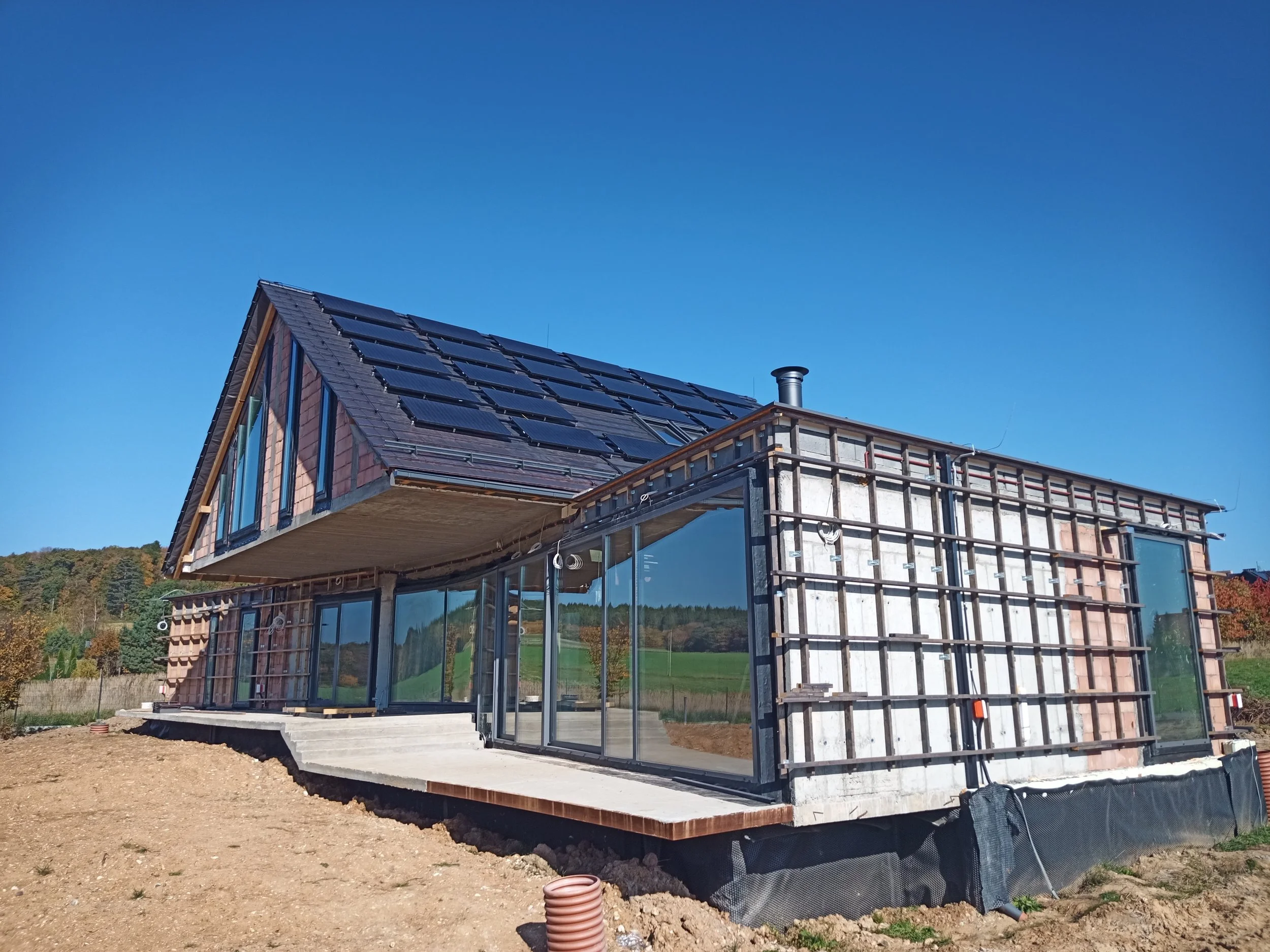 Under construction modern house with solar panels on roof, large glass windows, and exposed framing, with a foundation and surrounding land in progress.