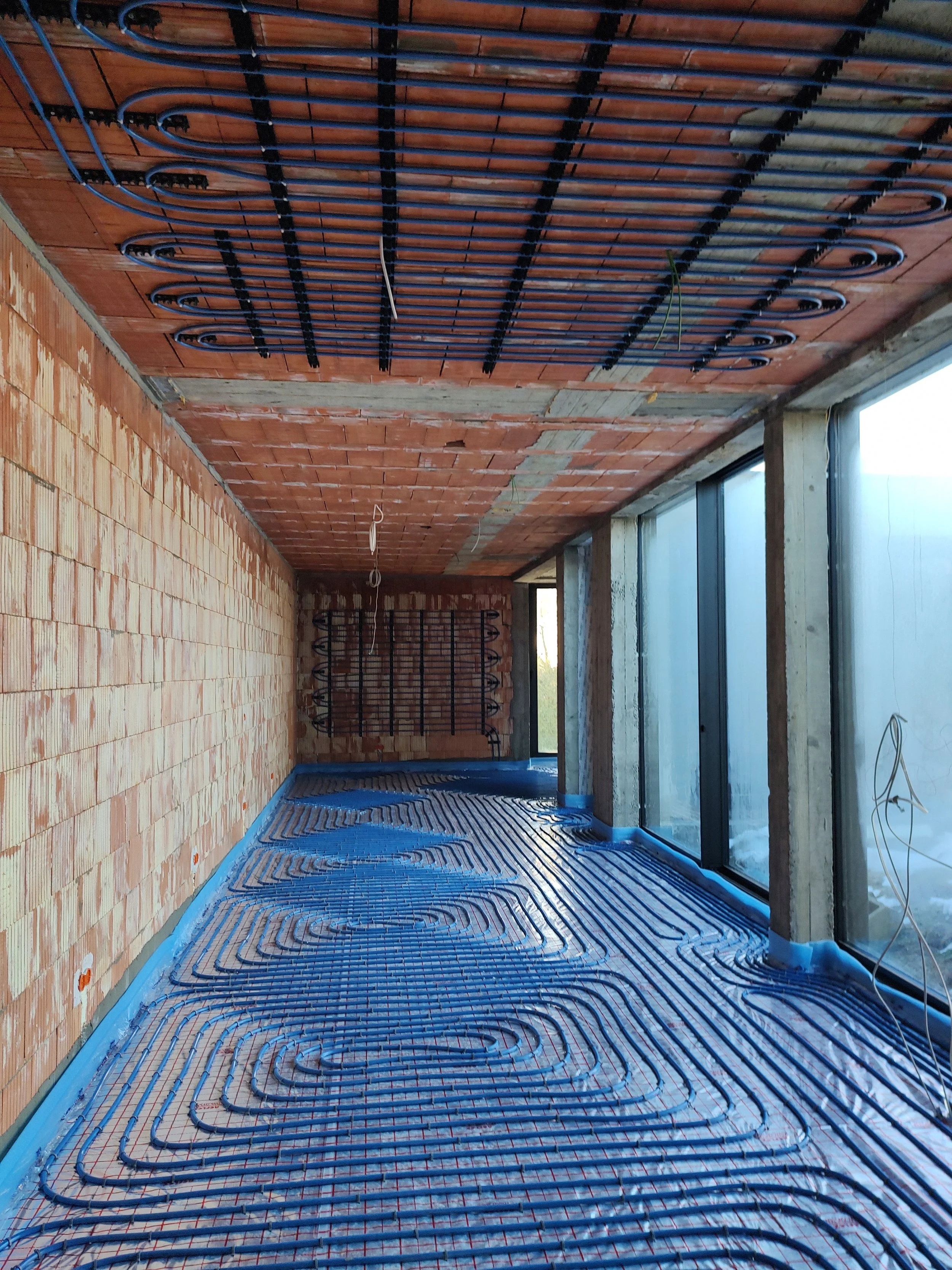 Under construction room with radiant floor heating system installed, exposed brick walls, large windows, and various wires hanging from the ceiling.