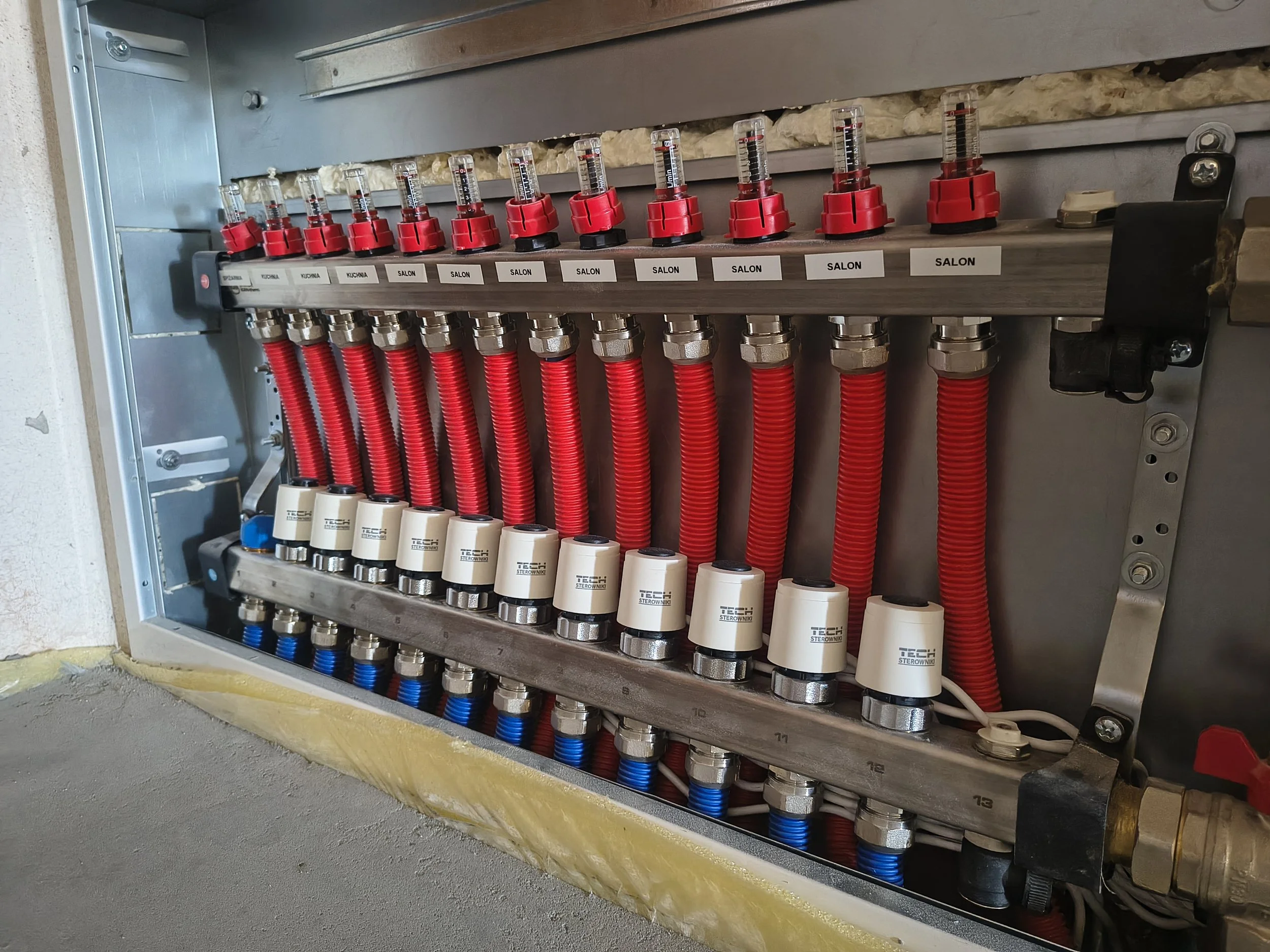 A mechanical manifold with multiple red and silver valves, black labels, and red, blue, and clear tubes connected to it installed in a utility box on a wall.