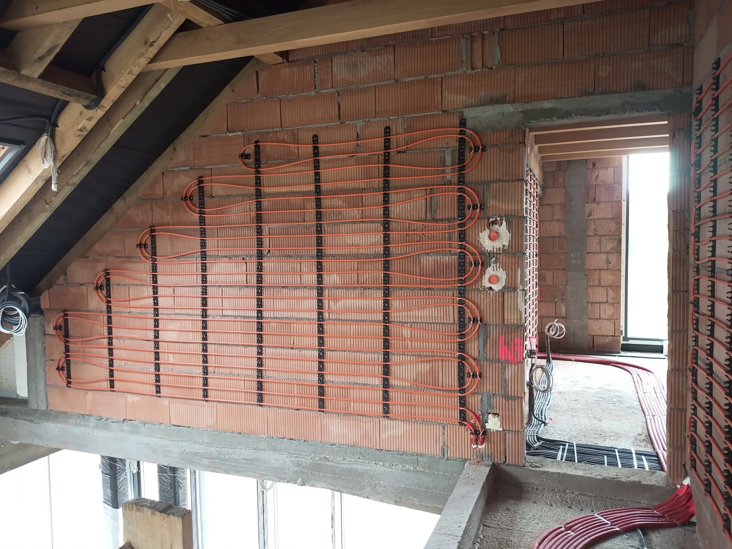 Interior of a building under construction showing an unfinished wall with orange in-floor radiant heating pipes and electrical wiring.