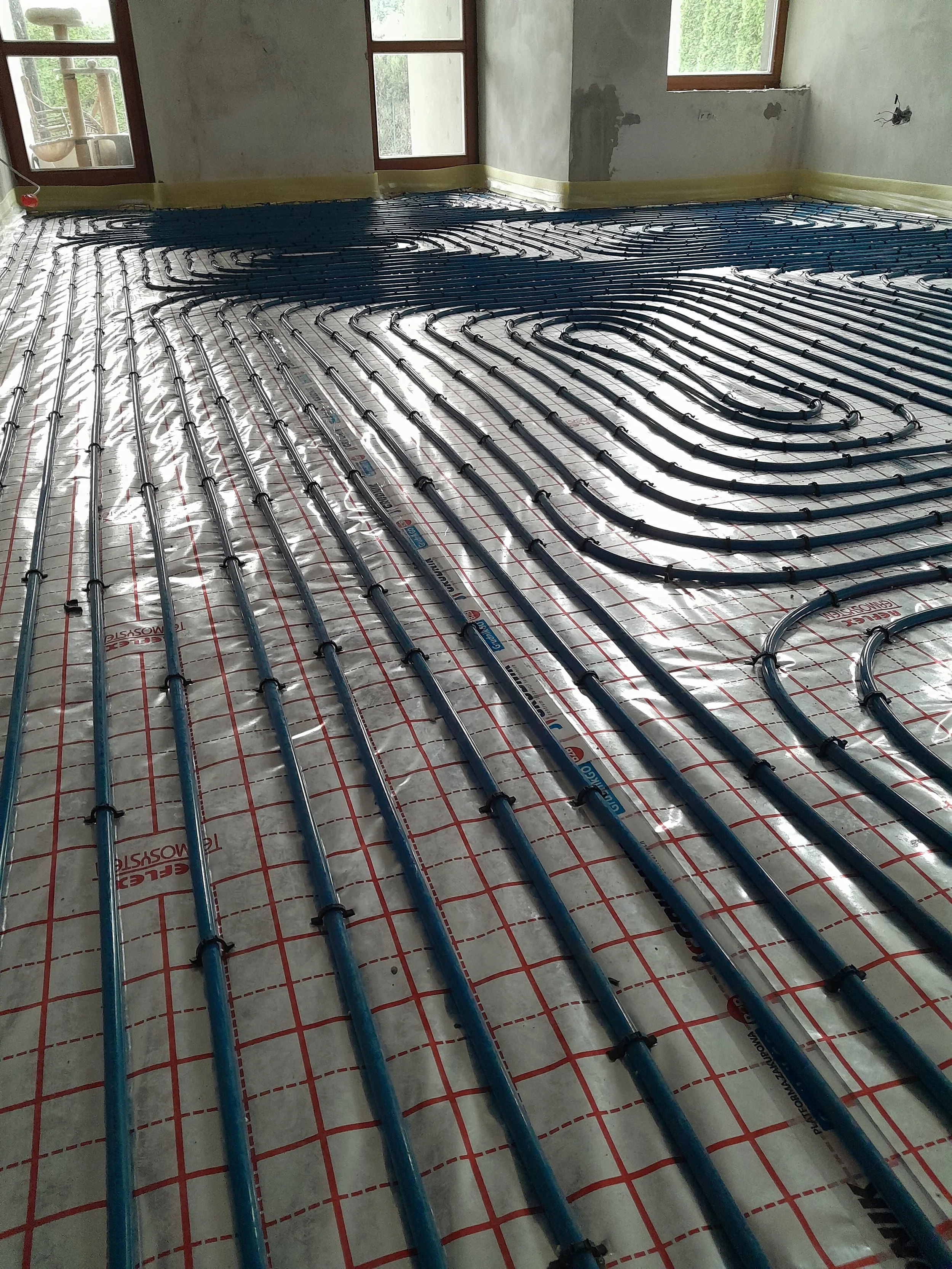A room under construction with an underfloor heating system installed. Blue pipes are laid out in a grid pattern across the floor, secured with black clips, on top of a reflective insulation layer with red grid lines. The room has three windows, some