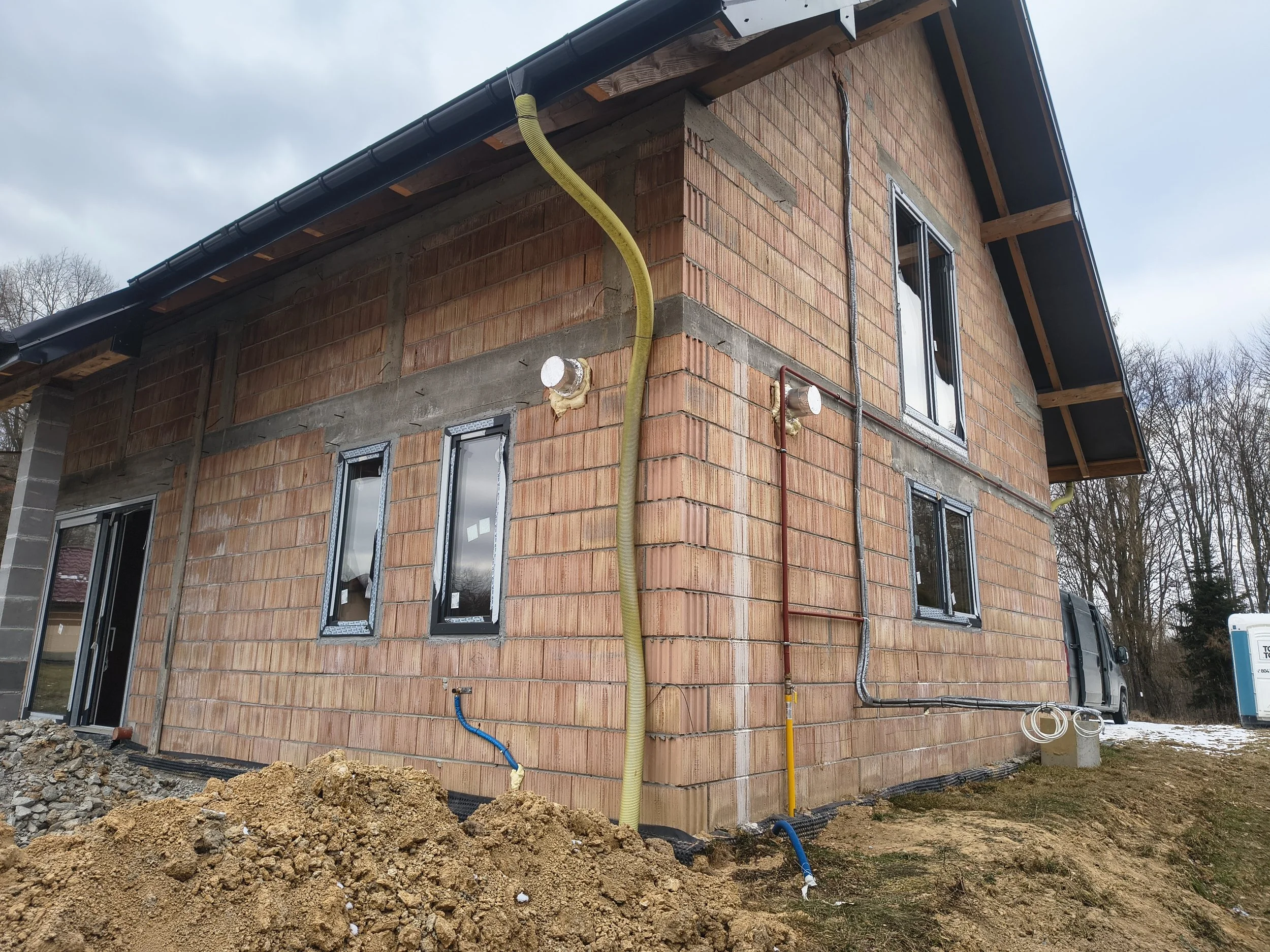 A house under construction with unfinished brick walls, multiple windows, and visible plumbing and electrical conduits on the exterior. There is a pile of dirt in the foreground and construction materials around the site.