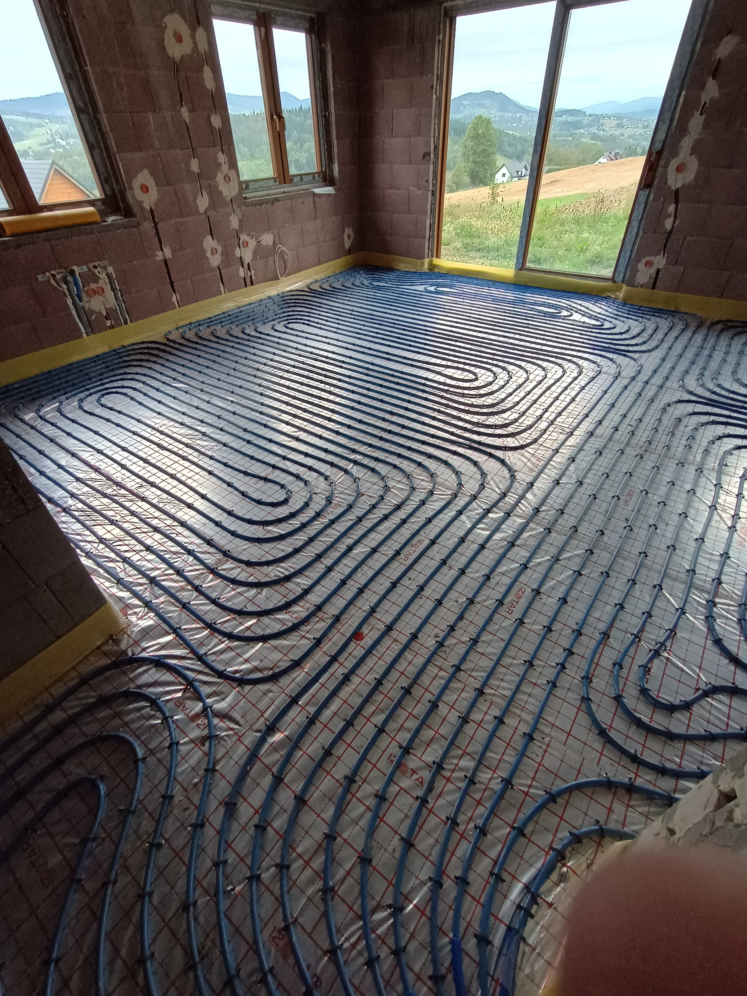 Room under construction with underfloor heating pipes installed, view of windows showing a mountainous landscape outside.