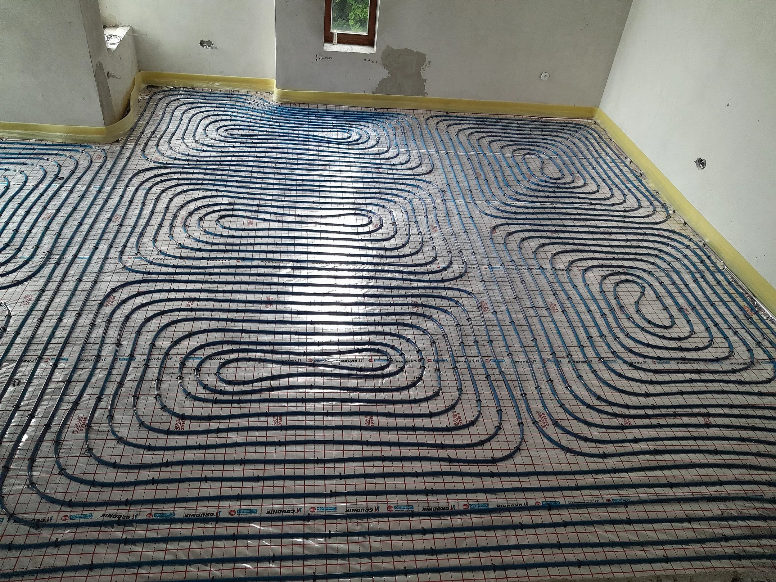 Underfloor heating pipes installed on a concrete floor in an unfinished room, with exposed walls and a single window.