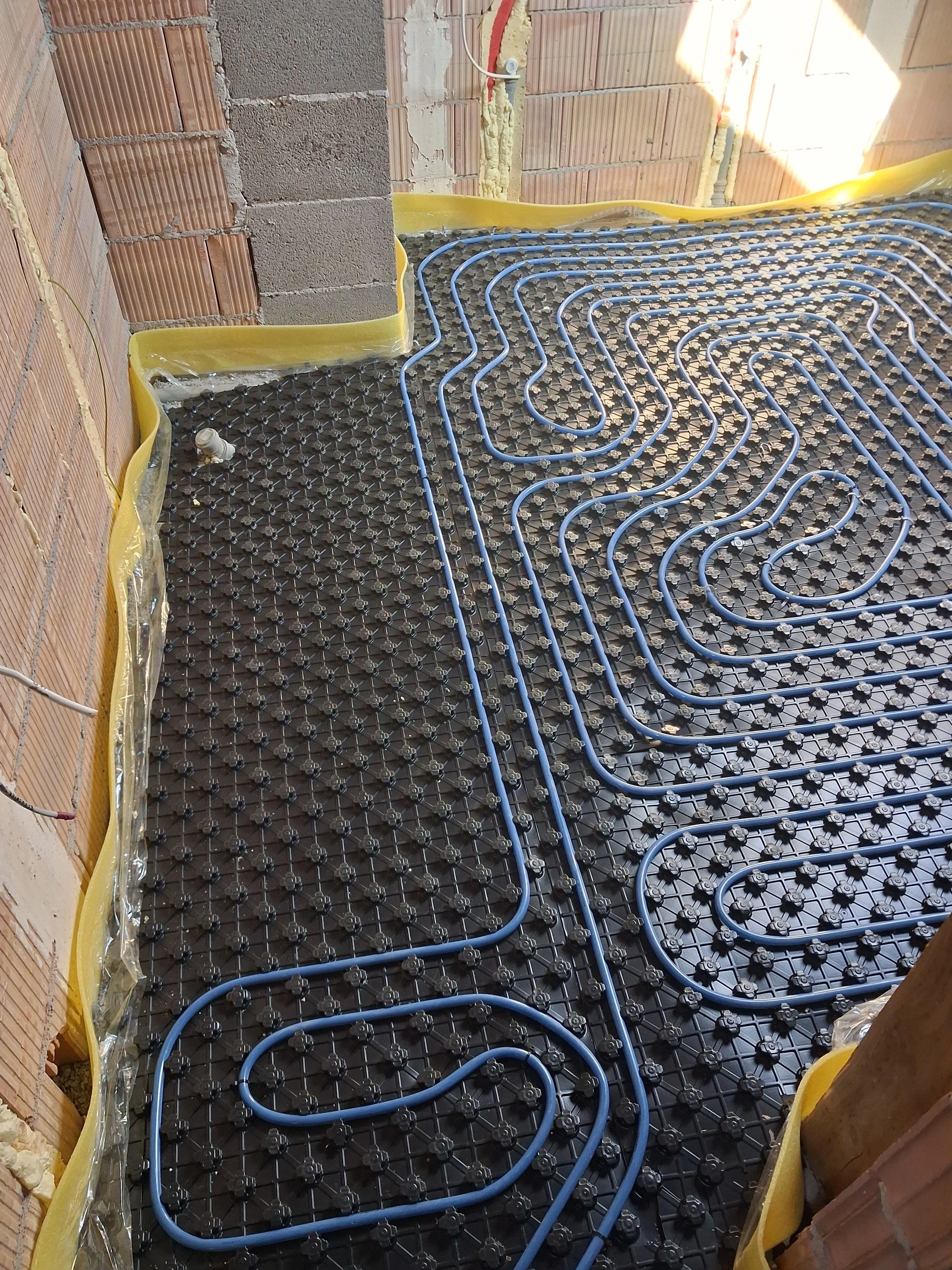 Underfloor radiant heating system installation with blue PEX tubing laid in a serpentine pattern on a black plastic grid in a room with brick walls and yellow waterproofing membrane.