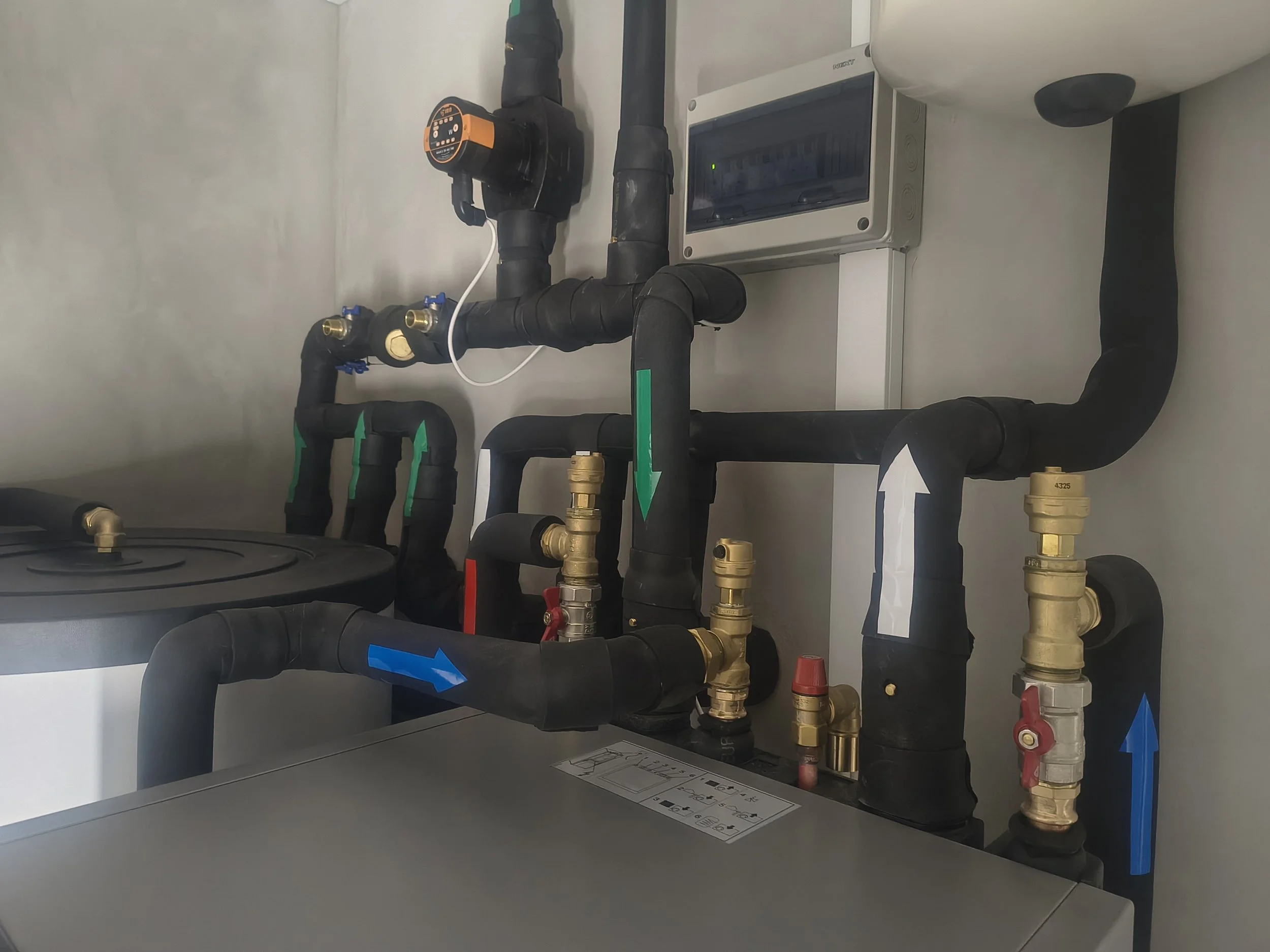 A network of black pipes with brass fittings and blue and red valve handles, connected to an electronic control panel, on a white wall.
