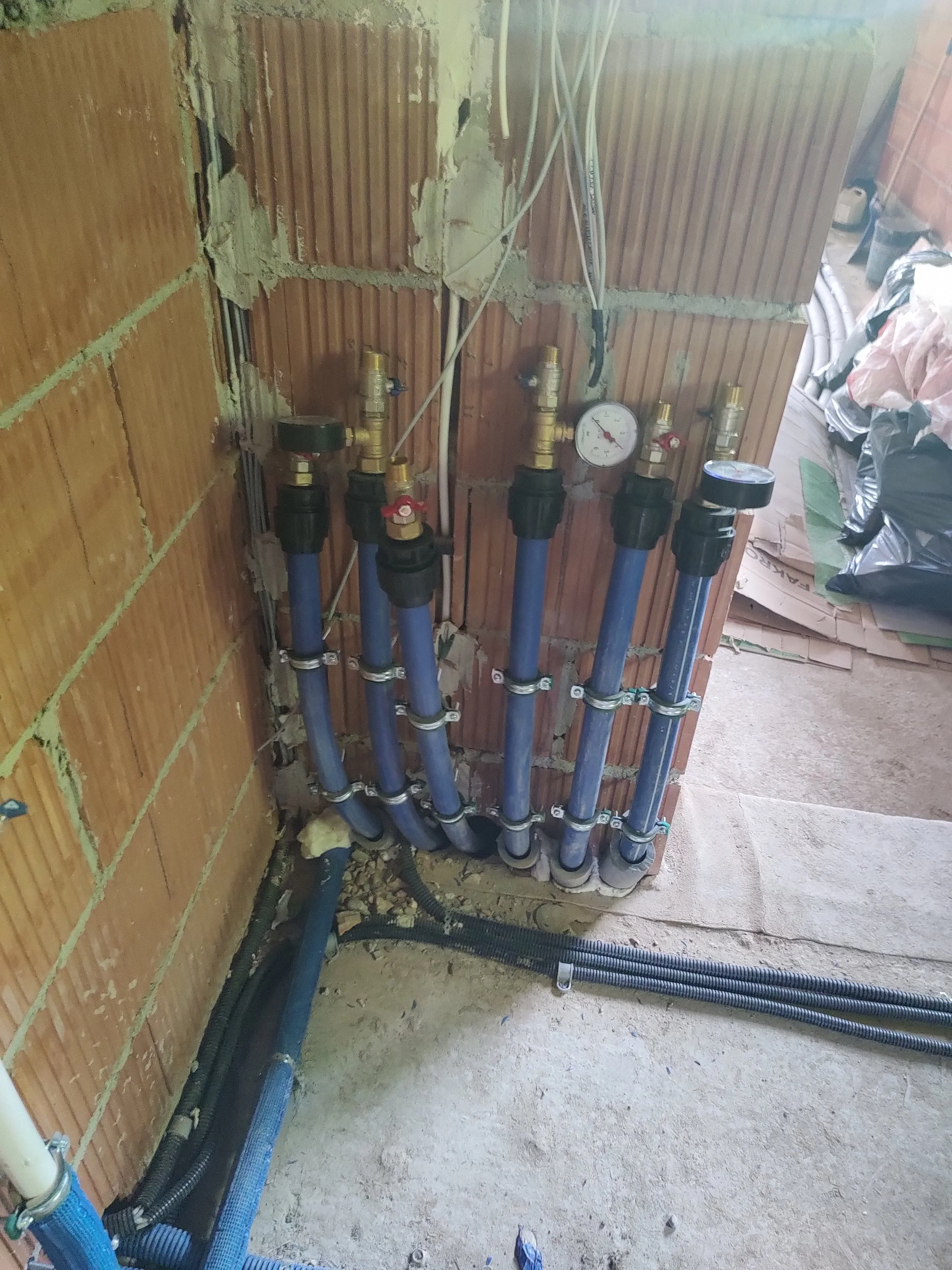 Water supply pipes, pressure gauge, and electrical wiring installed on a brick wall with unfinished sections and construction materials nearby.
