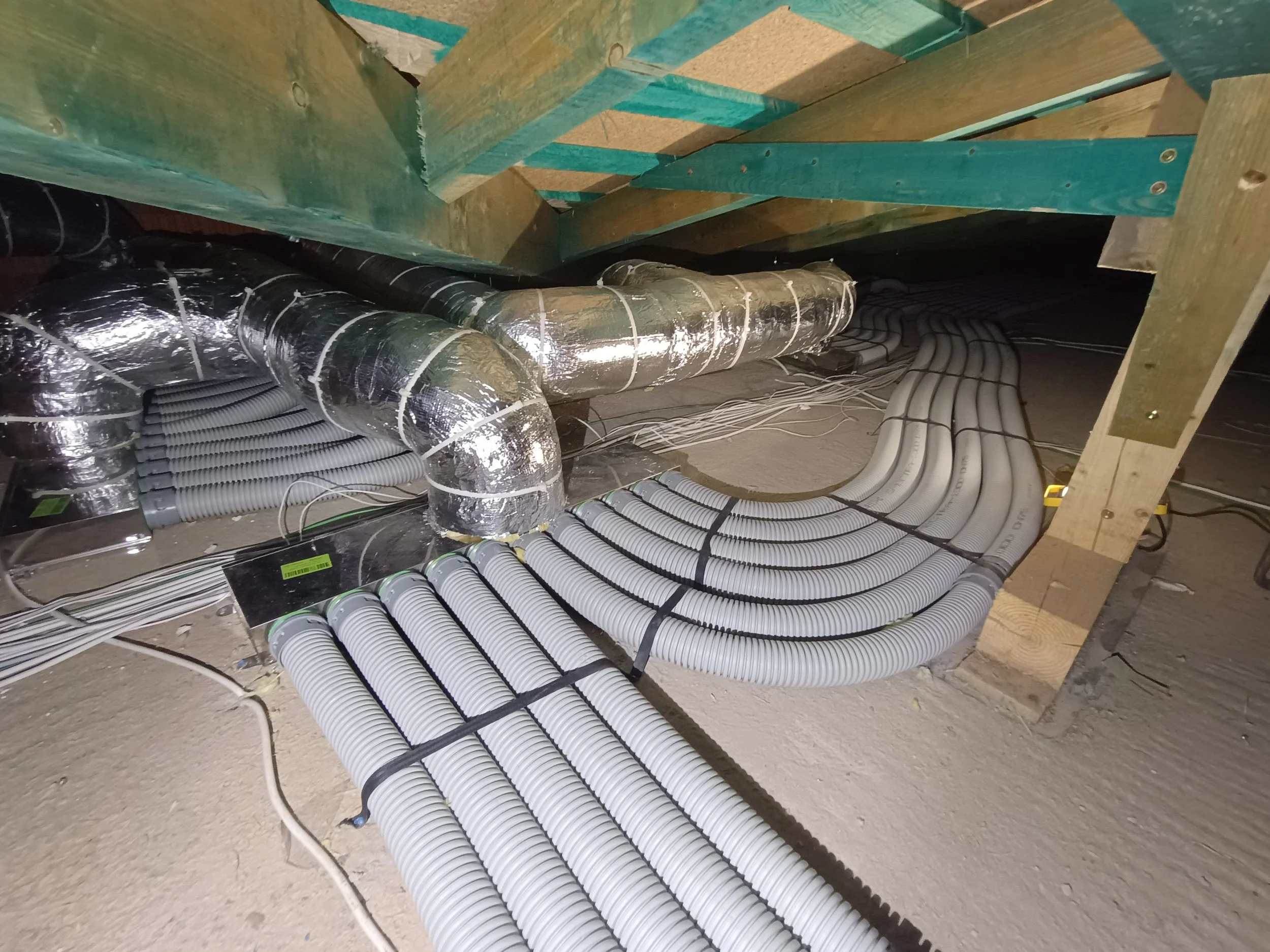 Underground utility installation with multiple white and black flexible conduits and insulated silver air ducts on a concrete floor, supported by wooden framing.