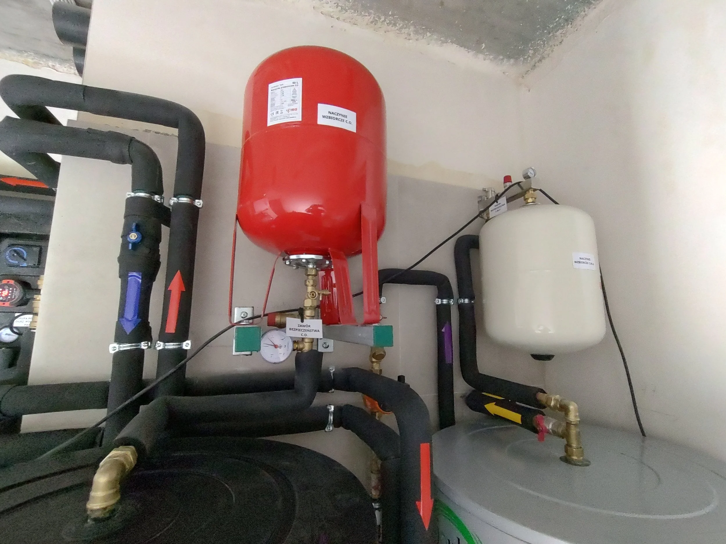 A fire safety system panel with pipes, valves, and gauges installed on a wall. The red tank has labels and is connected to black pipes with blue and red arrows indicating flow directions.