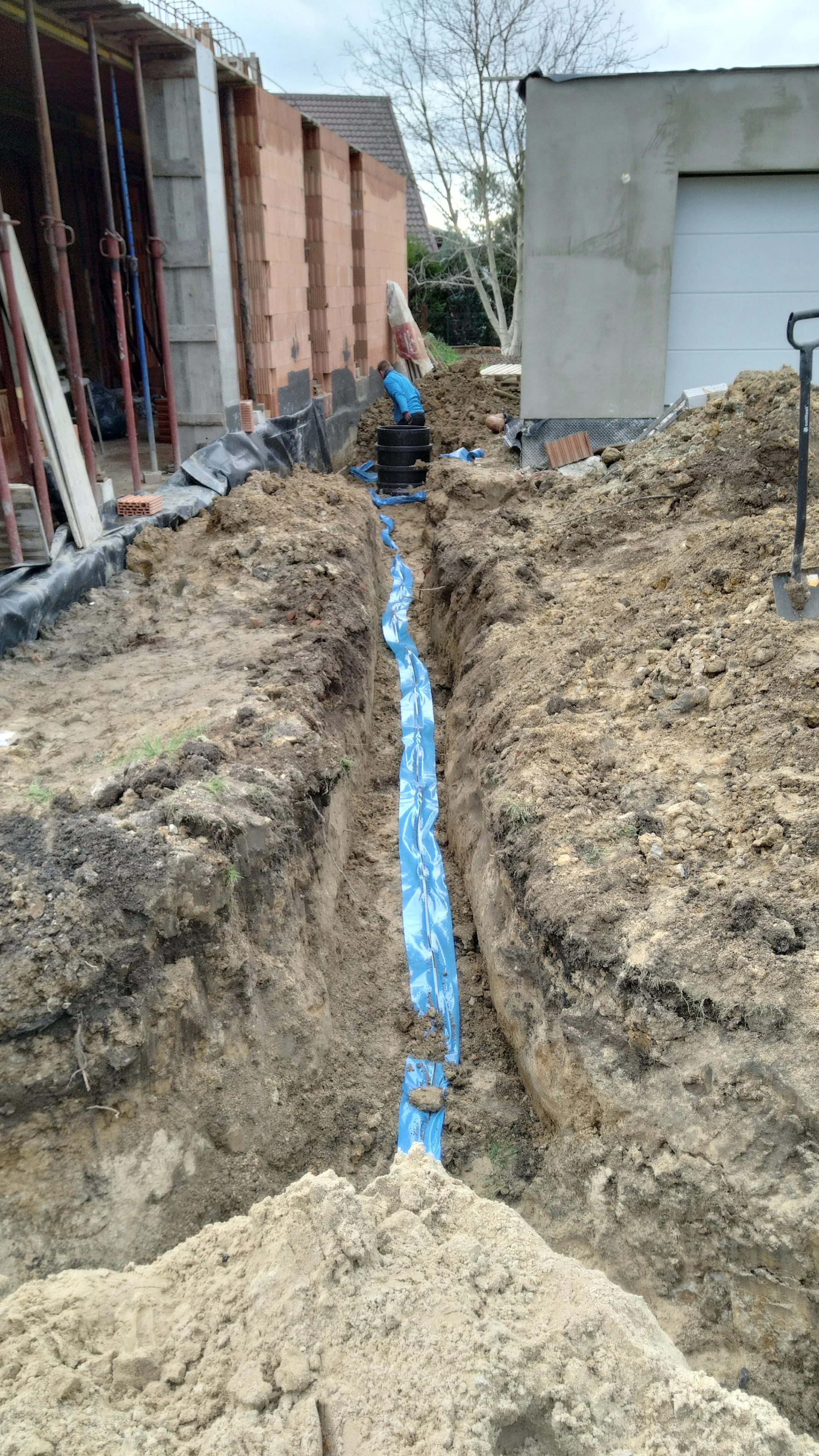 Construction site with a trench where a blue waterproofing membrane is being installed on the bottom. Two workers are working in the trench, one bending down and the other standing. Building foundations are under construction with bricks and concrete