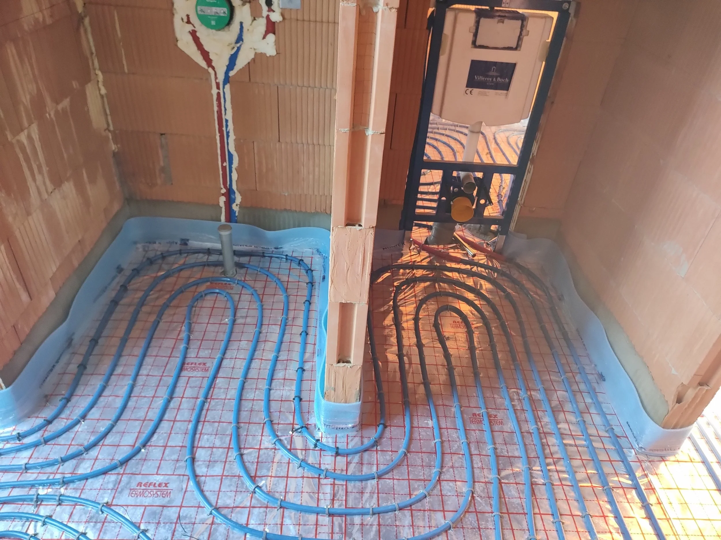 Installation of underfloor heating pipes on a wooden floor in a room under construction. The pipes are blue and laid out in a spiral pattern over insulation panels.