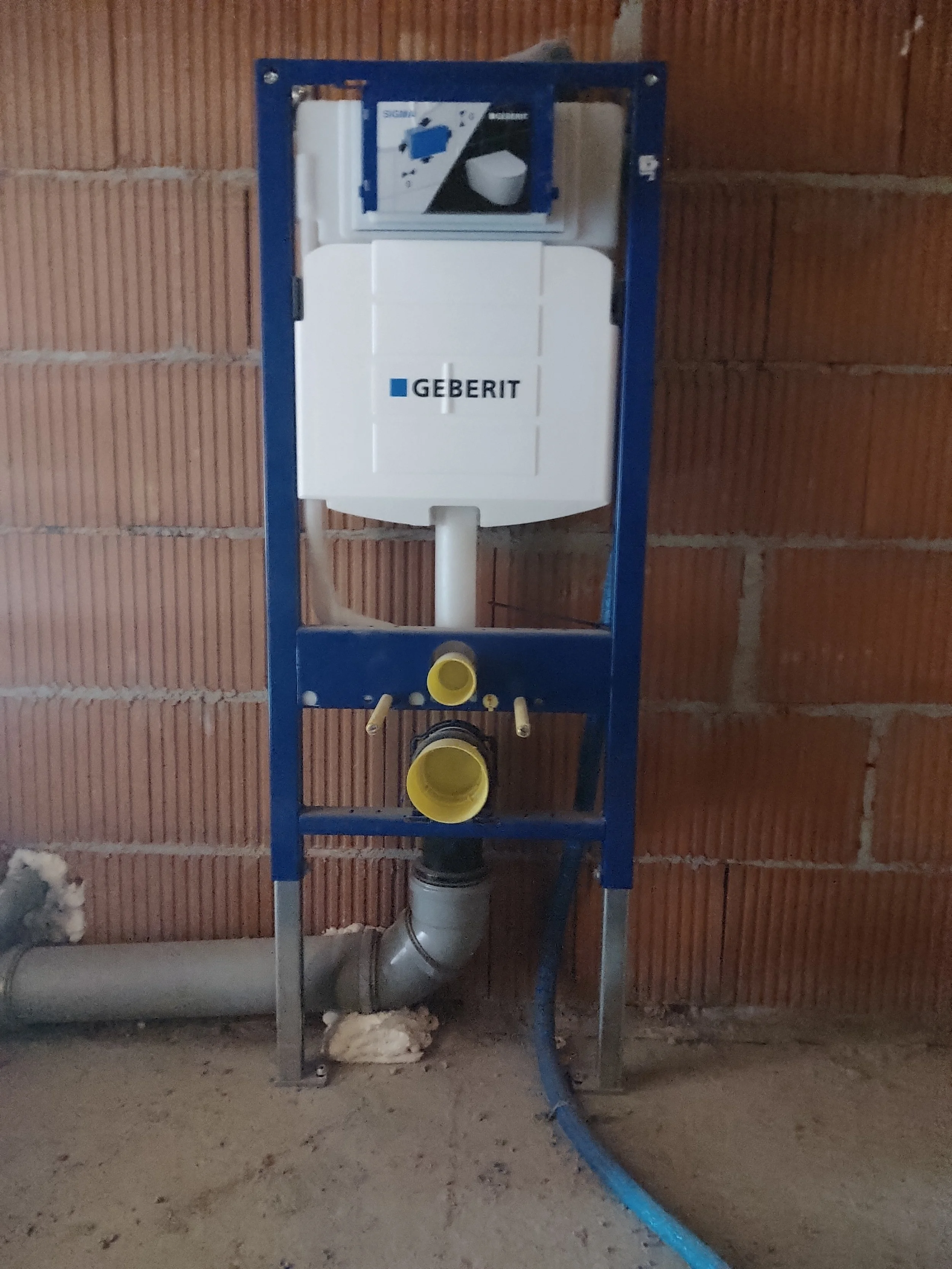 A partially installed toilet system with a white Geberit concealed cistern and yellow waste pipe, mounted on a blue metal frame against a brick wall, with a flexible piping connected to the waste pipe on a construction site.
