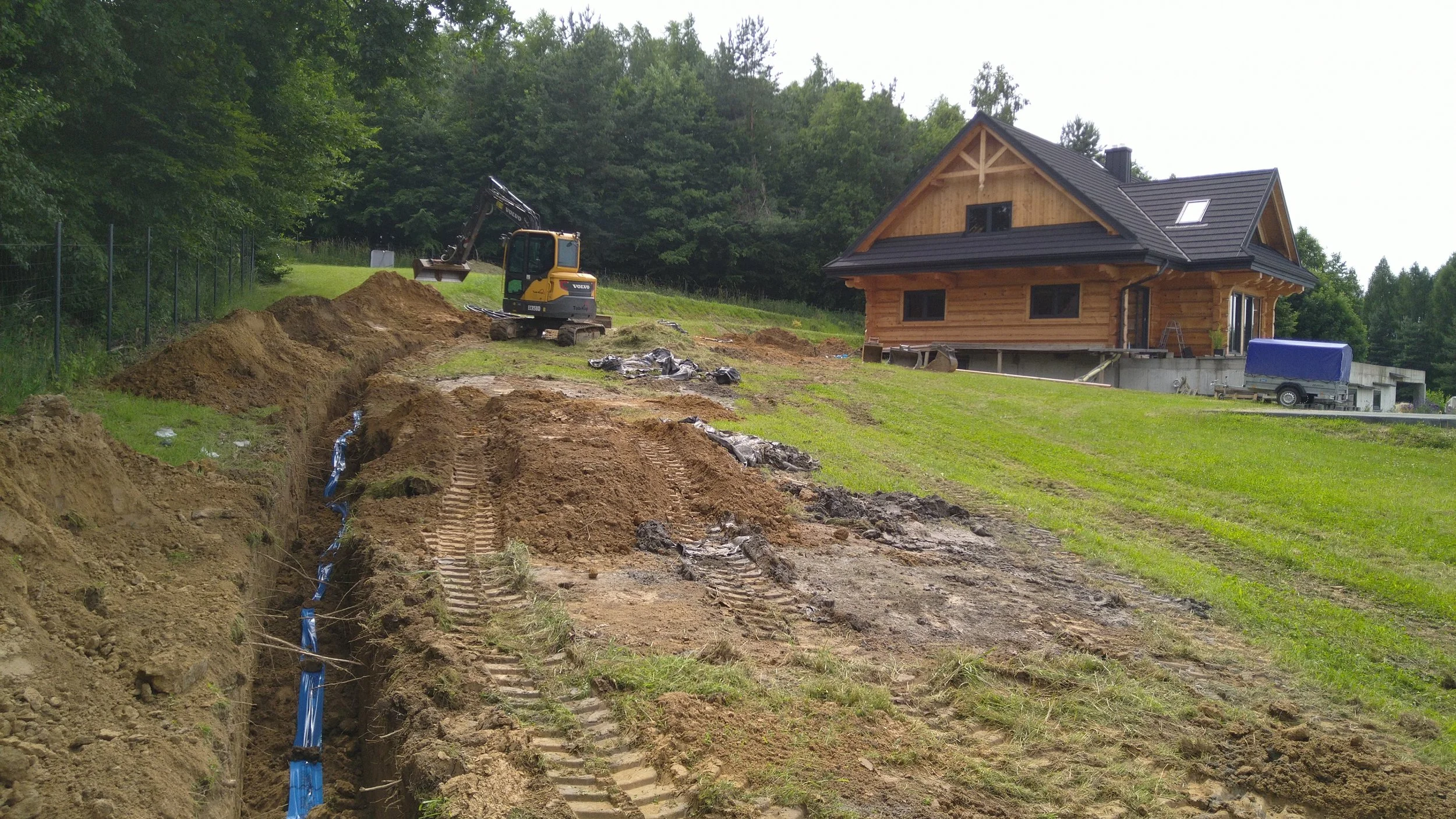 Construction site with a small excavator digging a trench, a planned house in the background, and a trailer on the right.