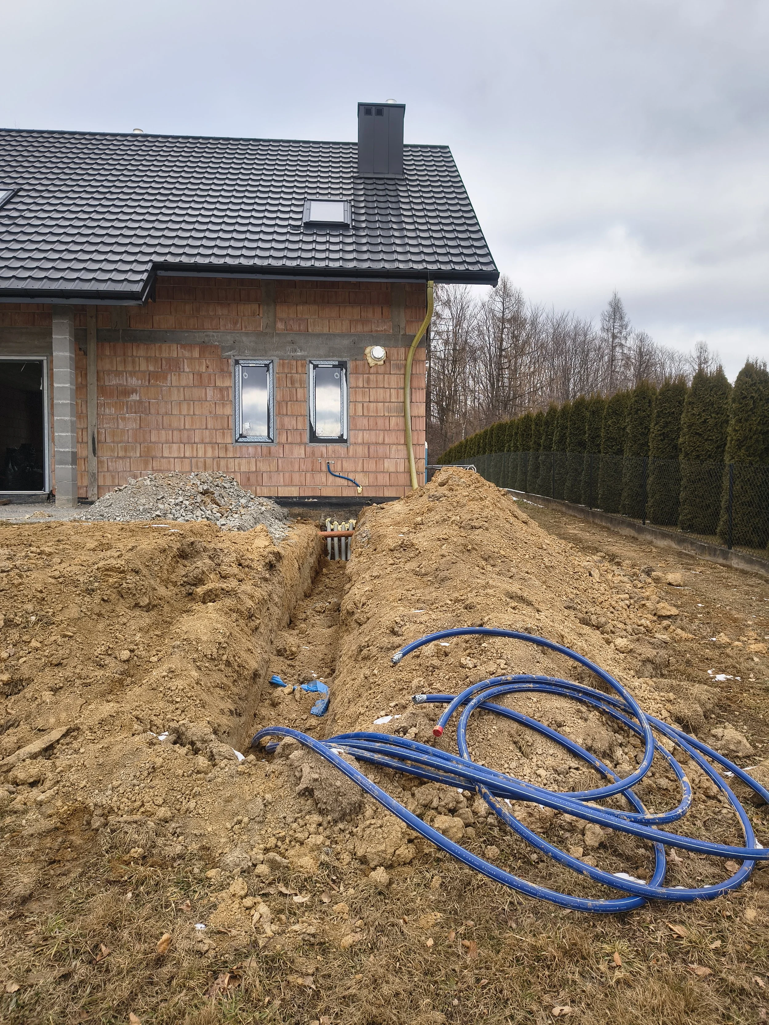 Construction site with an open trench and blue pipes, a house under construction with brick walls, windows, and a black roof, surrounded by dirt and some leafless trees.