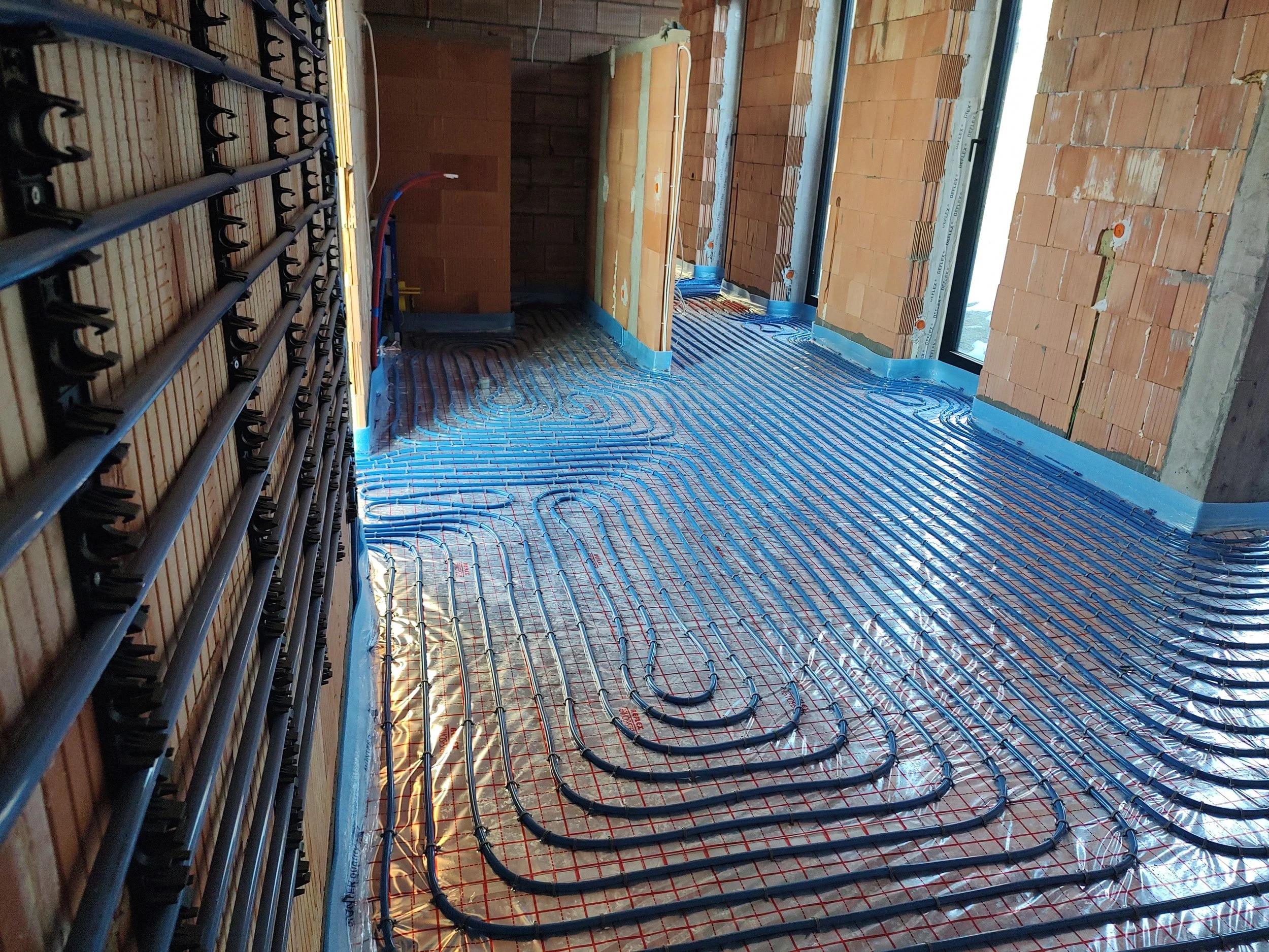 Construction site with underfloor heating being installed, showing heated pipes laid out in a pattern on the floor, with brick walls and window frames in the background.