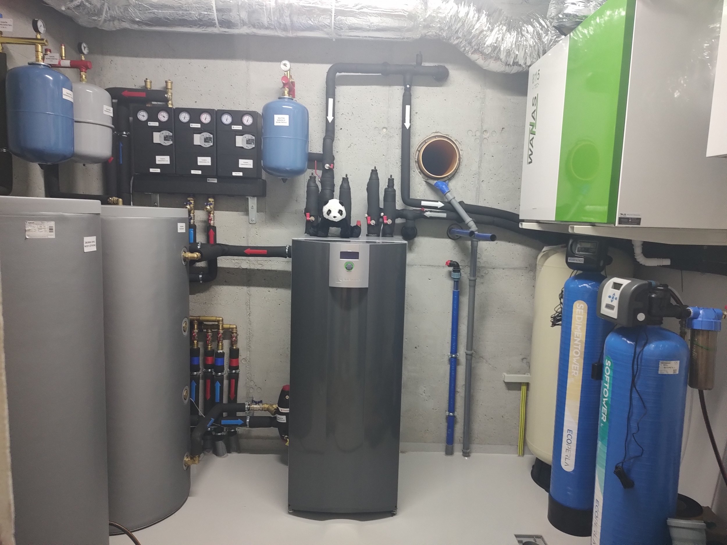 A utility room with various water filtration and heating equipment, including tanks, pipes, gauges, and a control unit, with a small panda plush toy placed on a gray box.