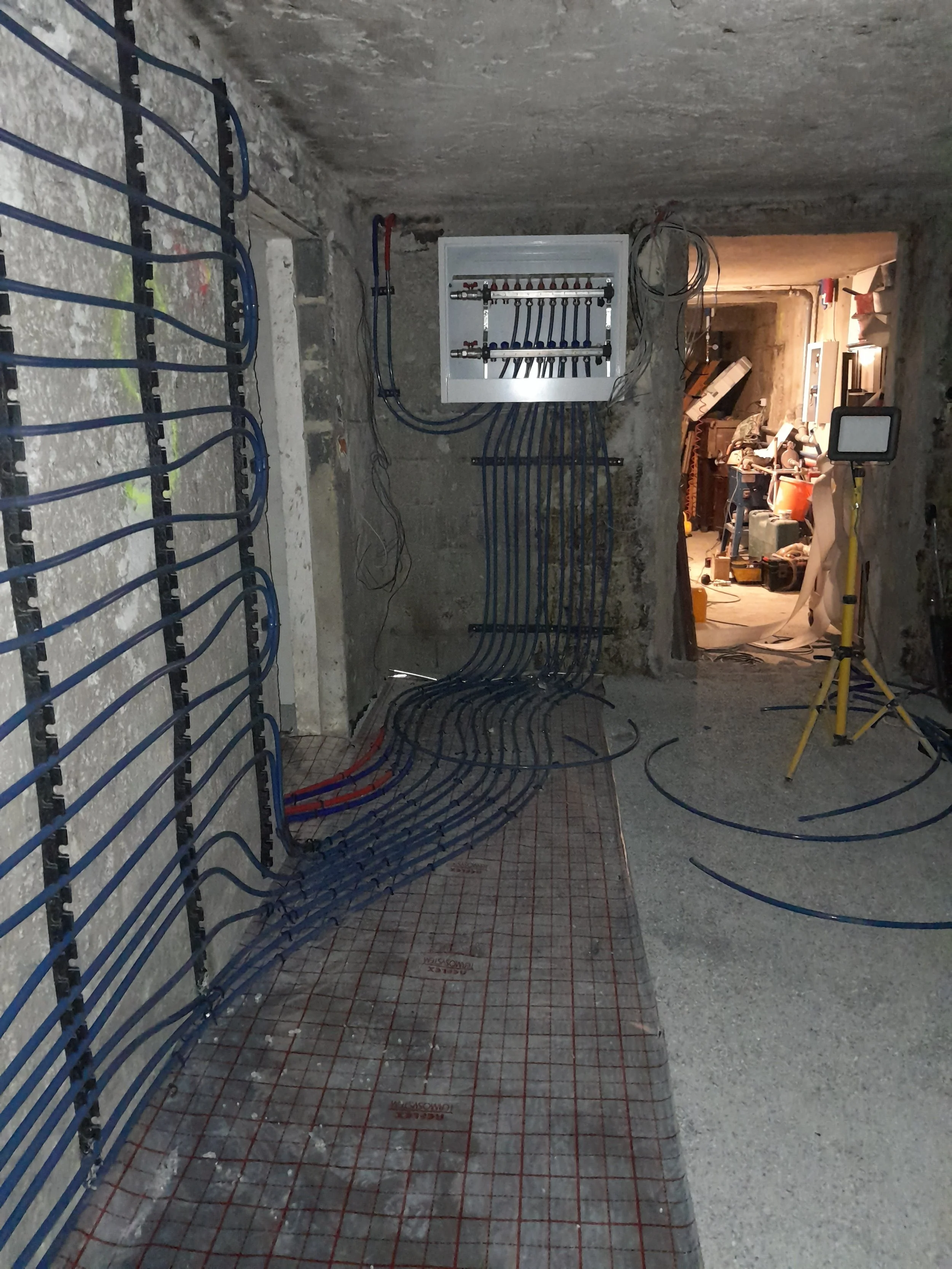 Construction site showing exposed blue electrical wiring and piping, with a concrete wall and door opening revealing tools and equipment inside.