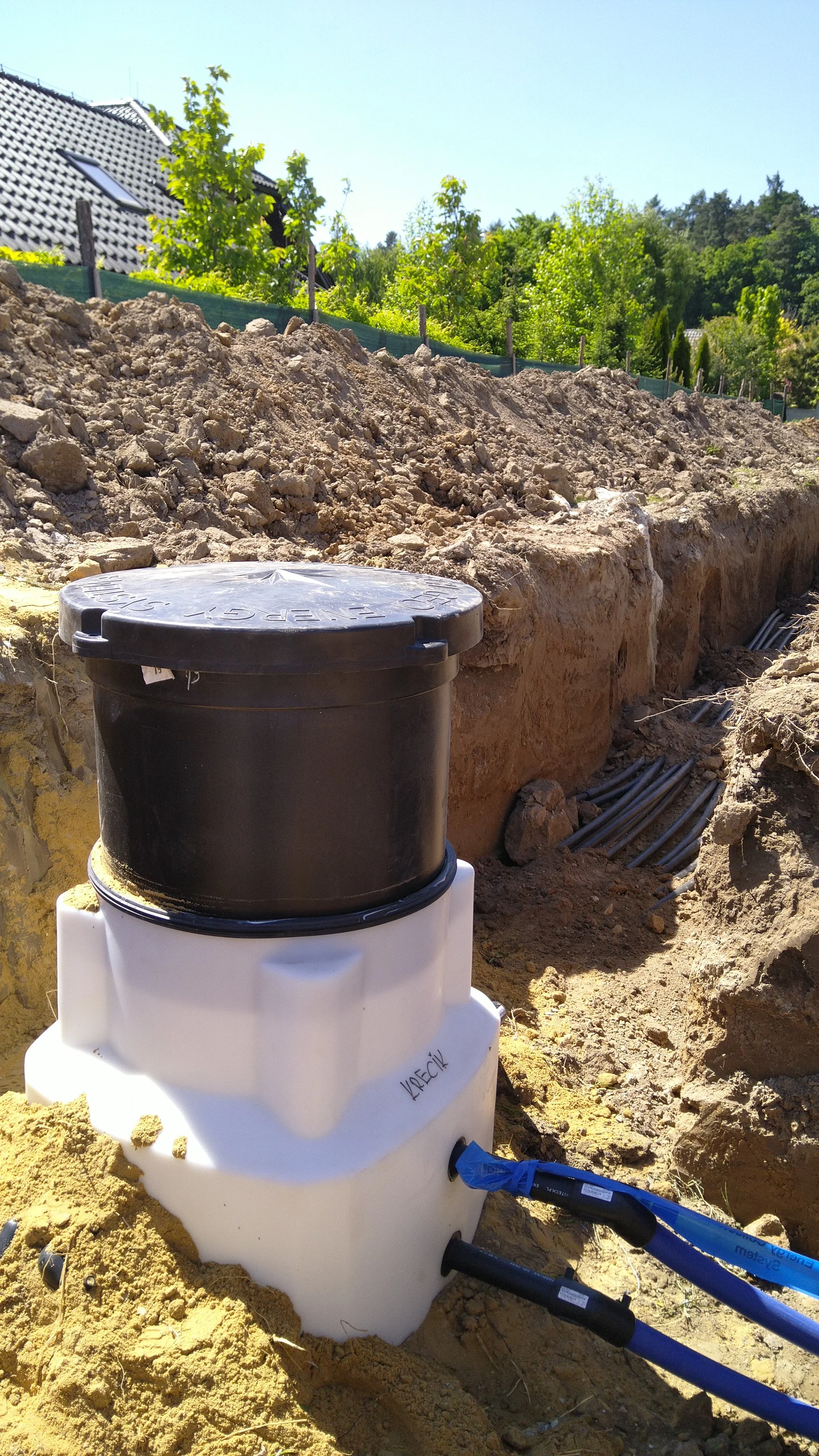 A septic tank installed in an excavated trench on a sunny site with dirt and construction pipes nearby, and a background of trees and a house.